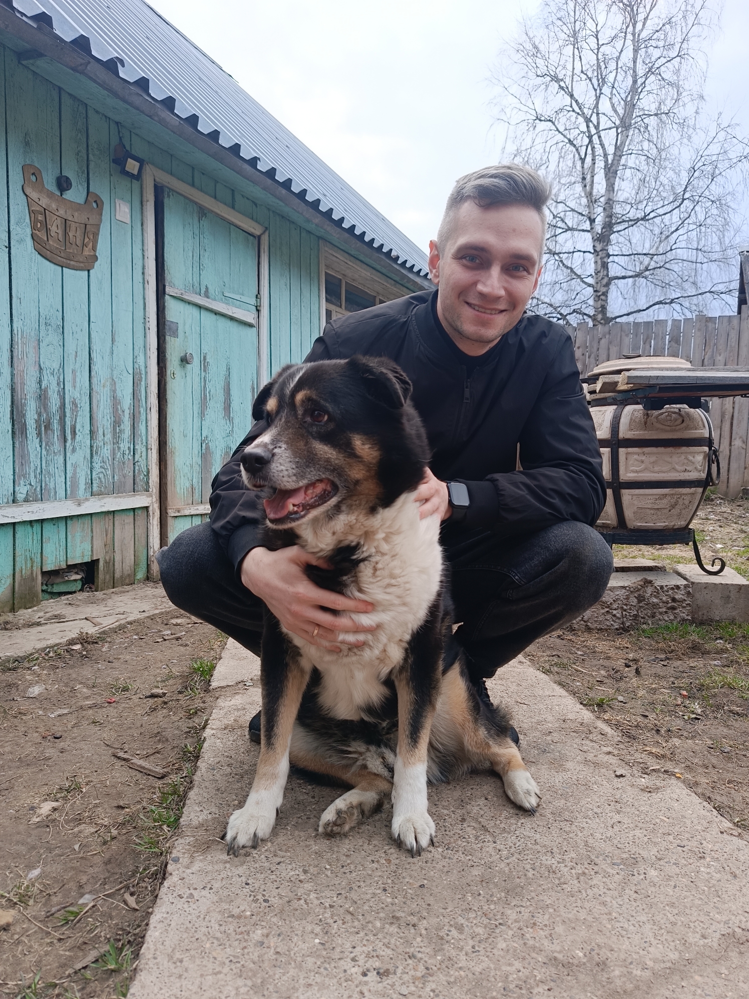 A man crouches beside a dog, posing for a photograph in what appears to be a rural or semi-rural setting in Rusalevka, Russia. The man, with short light-brown hair, wears a black bomber jacket and dark pants, and is smiling directly at the camera. His left hand rests gently on the dog's chest/shoulder. The dog, a medium-sized breed with a mix of black, white, and tan fur, sits upright with its mouth slightly ajar, looking contentedly to its right.
Behind them is a weathered light-blue wooden building with a dark corrugated metal roof, possibly a bathhouse or utility shed, given the wooden plaque hanging to the left of the door. This plaque is shaped like a wooden bucket and has the word "БАНЯ" (BANYA, meaning bathhouse or sauna) carved into it in Cyrillic. A bare deciduous tree stands tall in the background under an overcast sky, suggesting late winter or early spring. To the right of the man and dog, behind a low wooden fence, is a large, ornate ceramic or clay pot, likely a tandoor oven, resting on a metal stand and concrete blocks. The ground beneath them is a mix of dirt and patchy grass, with a concrete path running across the foreground where the man and dog are positioned. The overall atmosphere is casual and friendly, capturing a pleasant moment between a man and his dog.