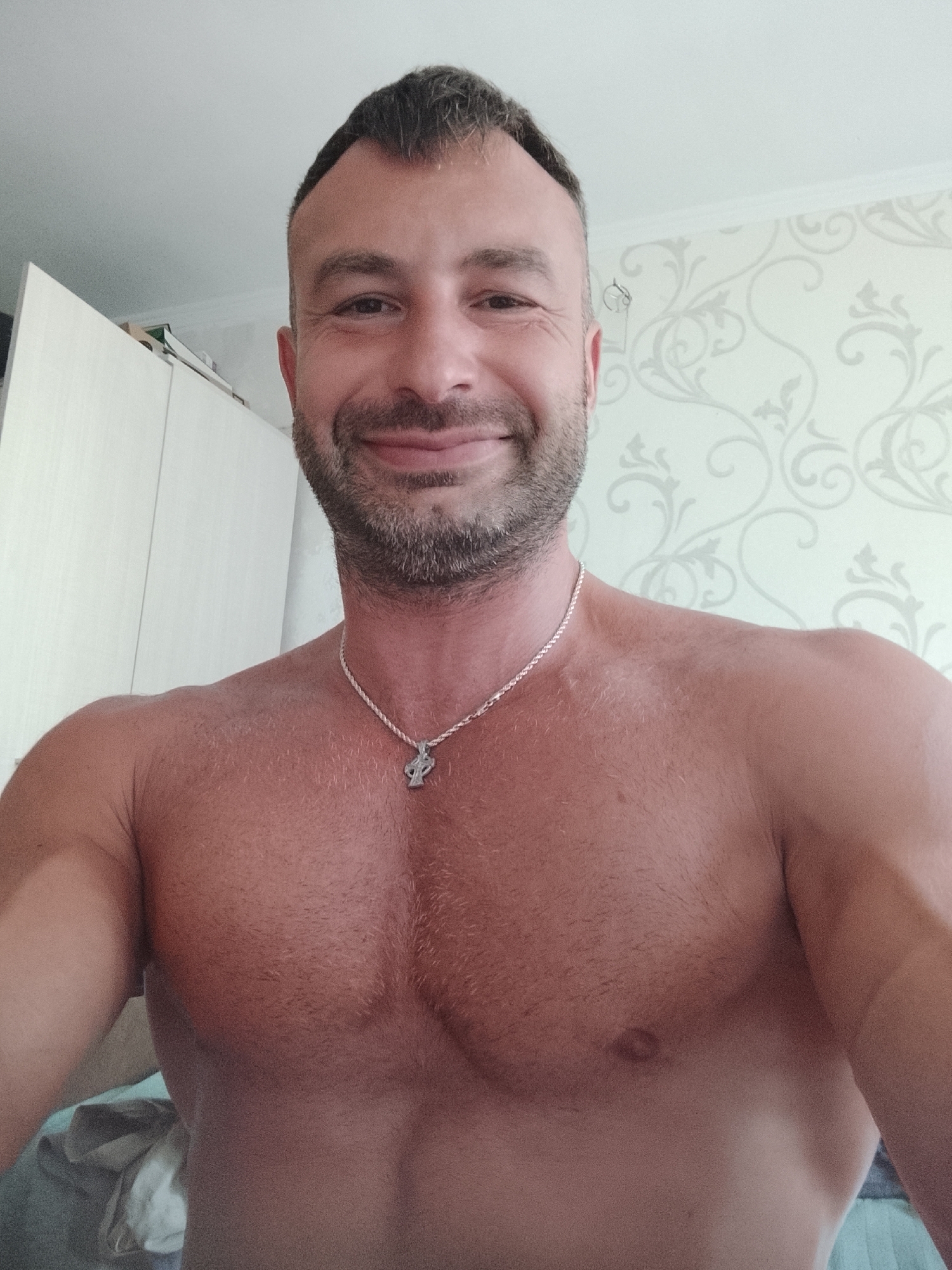 A man is posing for a selfie in a room. He is shirtless, wearing a silver necklace with a cross pendant. He has short, dark hair and a beard. The background shows a wall with patterned wallpaper and a closet. The lighting suggests it is daytime. There are no identifiable landmarks or clues to pinpoint the location to Chișinău, Moldova.