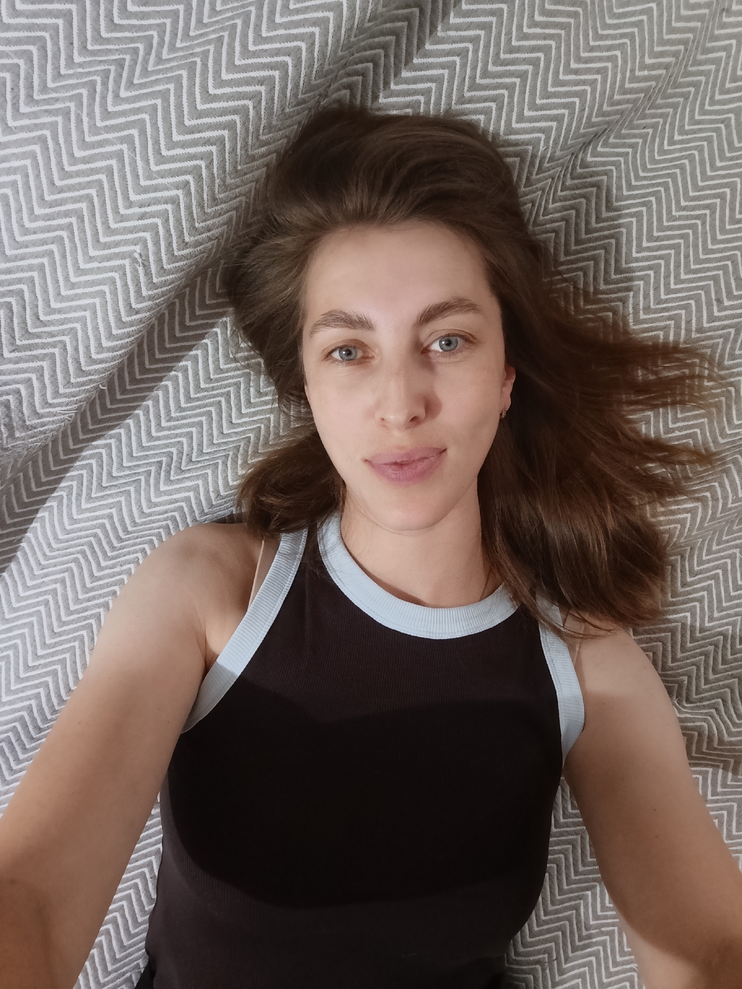 This is a selfie of a woman lying on a bed with a gray and white chevron-patterned blanket. She has long, brown hair that is spread out around her head. She is looking directly at the camera with blue eyes and a neutral expression. She is wearing a dark brown tank top with light blue trim. The lighting suggests it is daytime, likely indoors. There are no visible text or location cues besides the provided context of Chaikovsky, Russia.
