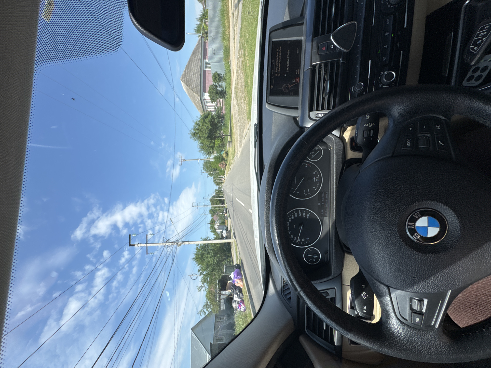 This is a first-person perspective video captured from the interior of a BMW car, likely in Mikhaylovsk, Russia, during the daytime. The scene is viewed through the windshield, which shows a road lined with houses and trees under a bright blue sky with scattered clouds. Power lines are prominently visible, crisscrossing the sky and poles.
The interior of the car reveals the steering wheel with the BMW logo, dashboard with a speedometer and other gauges, and a central infotainment screen displaying navigation and audio information. The text on the screen includes "Tvoy Durman feat. M," "Неизвестный альбом," and "ЗАКРЫВАЙ ГЛАЗА," suggesting music playback. The time is indicated as 9:39.
Outside the car, houses with distinctive pitched roofs and a roadside area with some discarded items, possibly household waste or donations, are visible. The overall impression is a drive through a residential area on a sunny day. The driver's hands are on the steering wheel, suggesting the car is in motion.