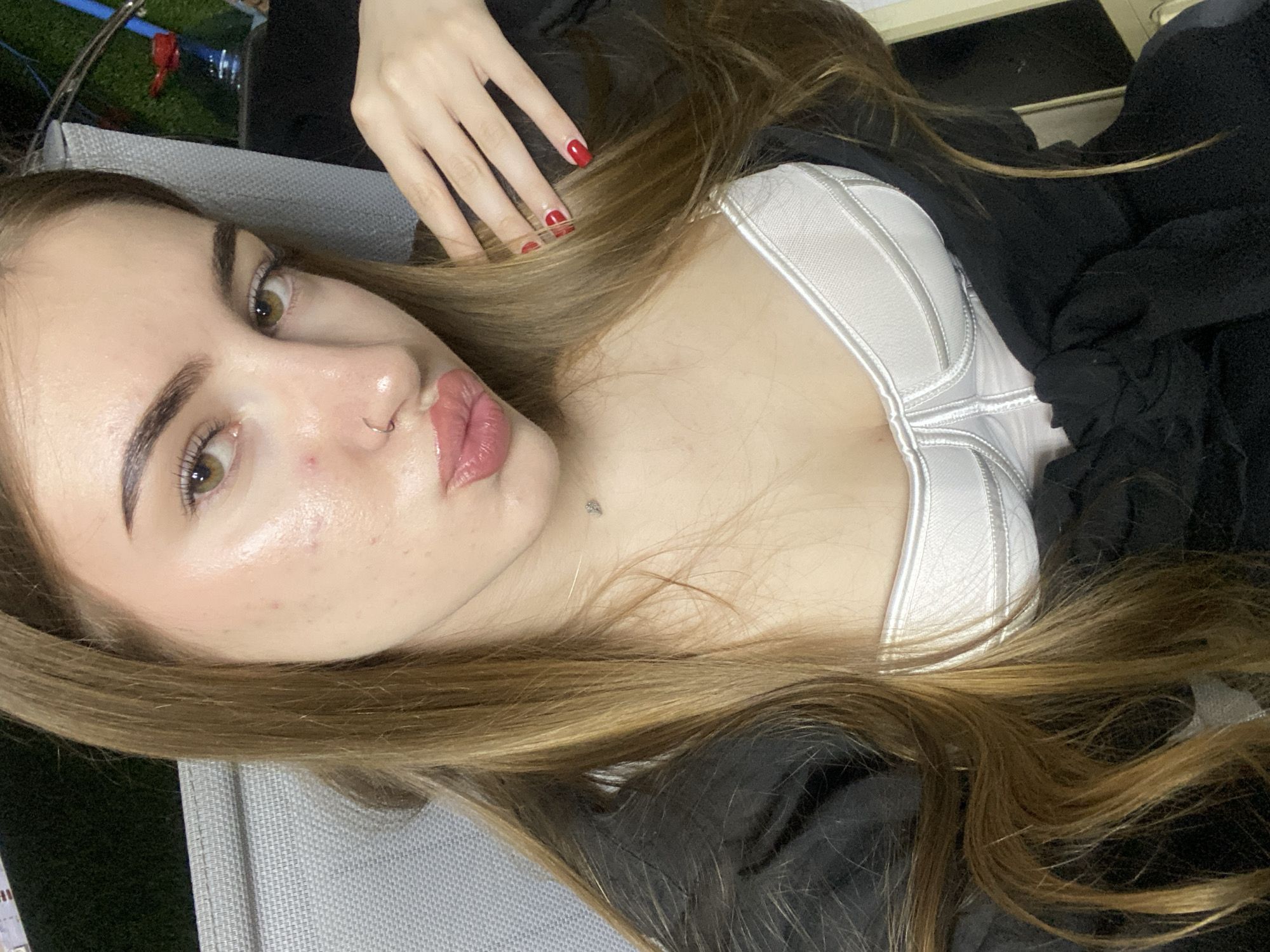 A young woman with long, blonde-brown hair and light brown eyes is posing for a selfie. She is wearing a white corset-style top and a black garment that appears to be a jacket or robe. Her lips are pursed, and she has a nose ring and a small mole on her neck. Her fingernails are painted red. The background is blurred, but suggests an outdoor setting with some furniture and greenery, possibly a patio. The lighting is warm and suggests it might be daytime. The image does not provide any specific details to identify a location in the city of Дьяково, Russia.