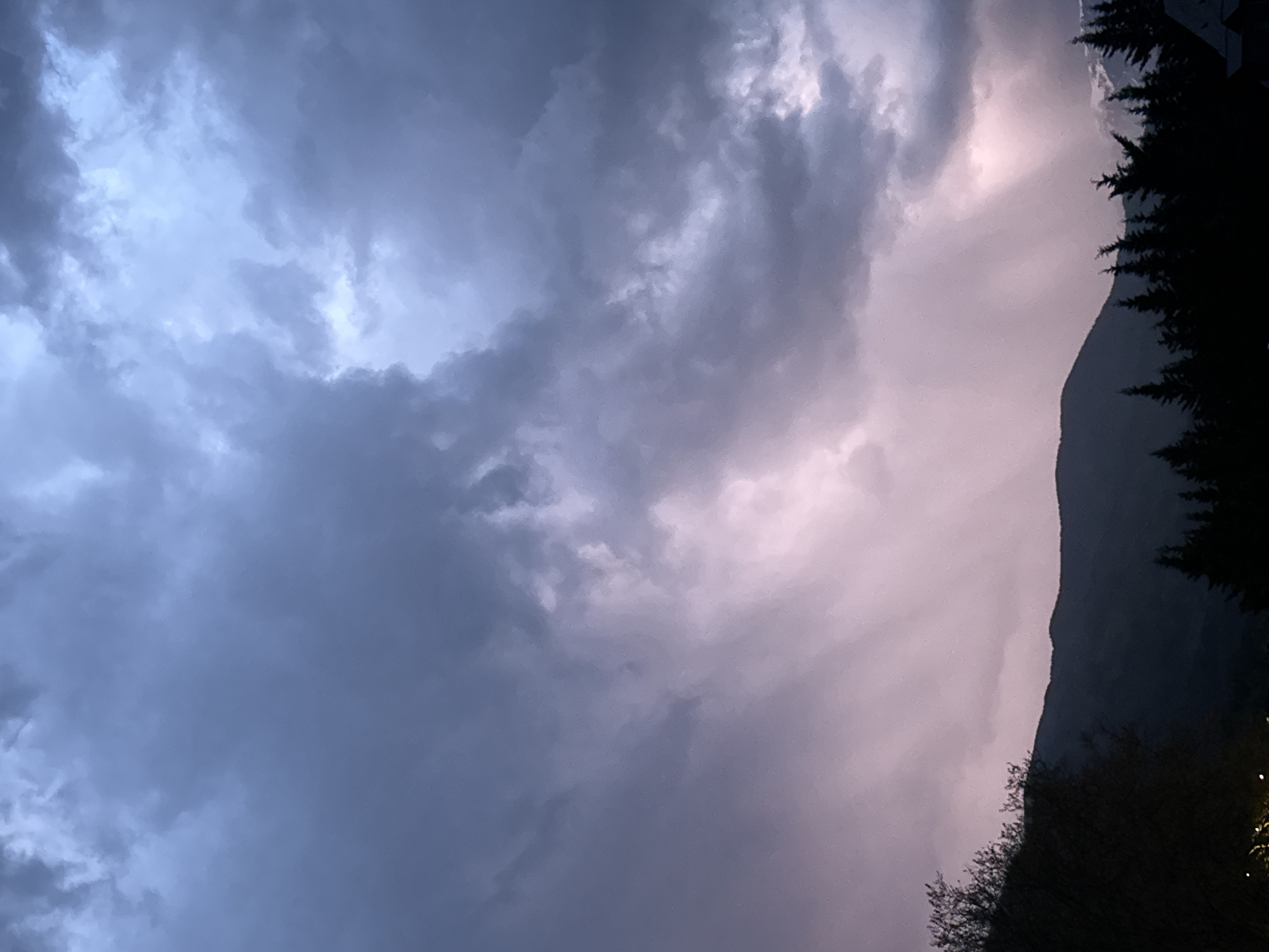 The image, oriented correctly, displays a dramatic and heavily clouded sky over a mountainous landscape, likely captured during dawn, dusk, or a stormy period. The sky is dominated by turbulent, textured clouds, ranging in color from deep bluish-grey on the left to a lighter, ethereal pinkish-white on the right, where a strong light source appears to be breaking through or reflecting off the clouds. This illumination creates a striking contrast and rays of light within the cloud formations on the right.
Along the right side of the frame, the dark silhouette of a rugged mountain range rises, its peak visible against the lighter sky. In the bottom right foreground, a dense cluster of dark trees, appearing to be conifers, adds to the shadowed, dramatic feel of the scene. The overall impression is one of immense natural power and grandeur, with the potential for changing weather. No human activity or visible text is present. The mountainous setting is consistent with the provided context of Benasque, Spain, located in the Pyrenees.