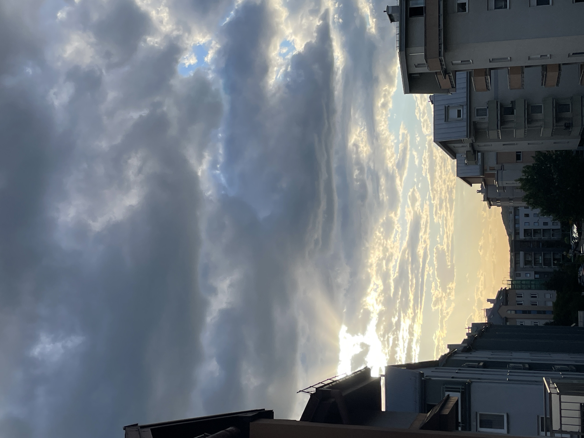 The image shows a view of buildings and sky, likely in an urban environment. The buildings are multi-story, with a mix of residential and possibly commercial structures visible. The sky is filled with clouds, some dark and heavy, others lighter and illuminated by sunlight. The sun appears to be setting or rising, casting a warm glow on the clouds and creating a dramatic contrast between light and shadow. The image is taken from a low angle, likely from a balcony or rooftop, looking up and out towards the horizon. The city is unidentified as Unknown. The image does not contain any people, actions or interactions.
