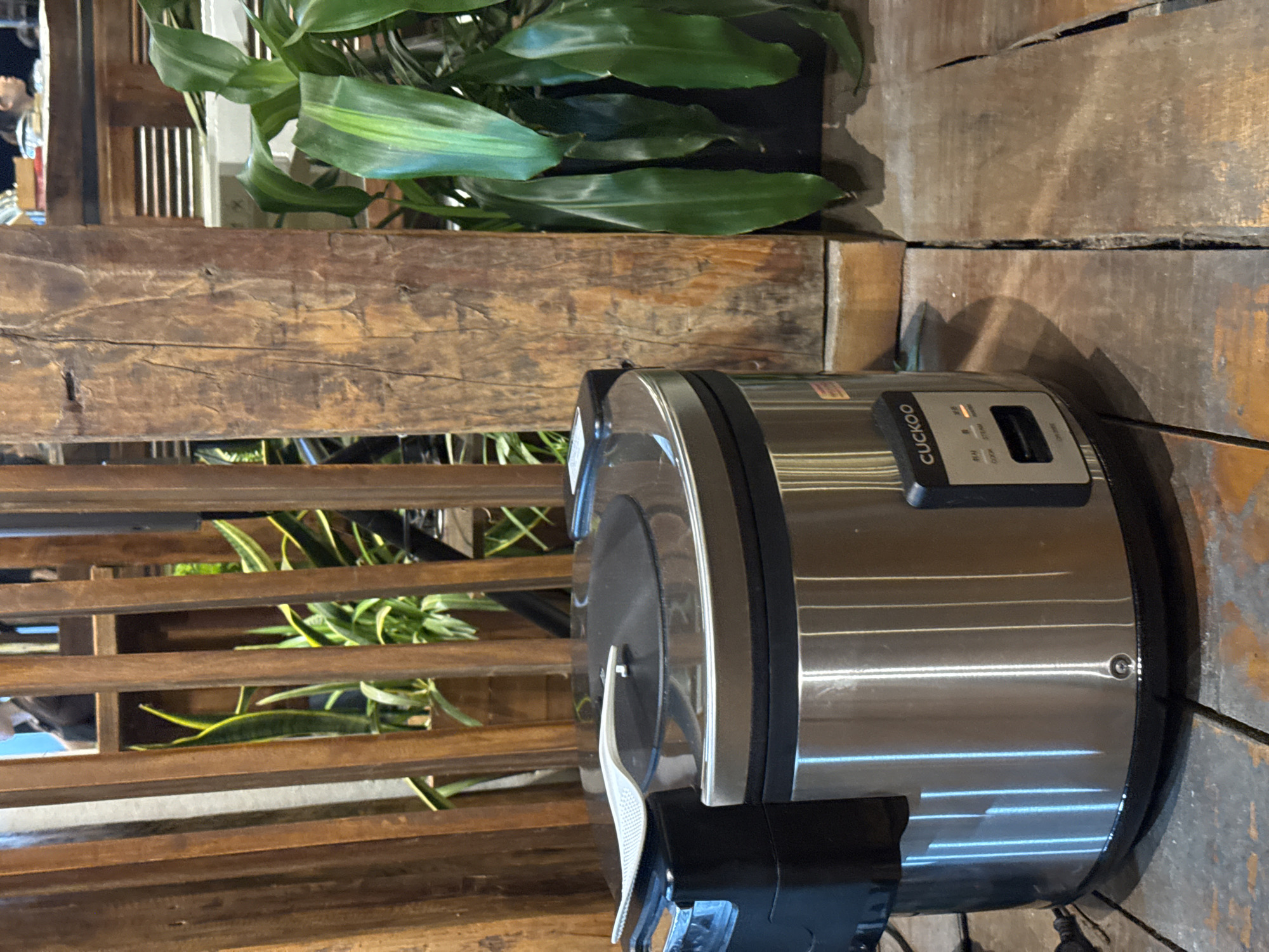 The image shows a silver Cuckoo brand rice cooker sitting on a wooden surface. The rice cooker is positioned on the right side of the frame. To the left, there is a wooden structure with horizontal slats, through which green plants are visible. Above the rice cooker and partially behind the wooden structure is another potted plant with broad green leaves. The wooden surface underneath the rice cooker appears to be made of planks.