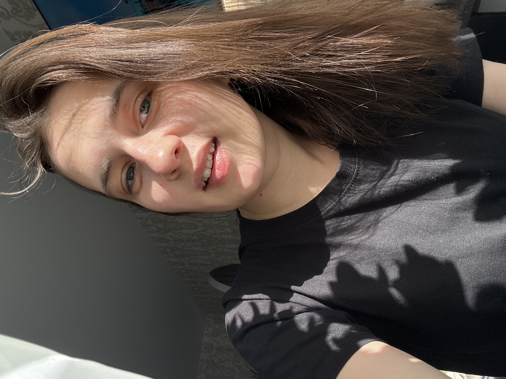 A young woman with long, straight, brown hair is the central subject of this close-up portrait, likely a selfie. She looks directly at the camera with a gentle, slight smile, revealing a hint of her teeth. Her light-colored eyes (blue/grey) are prominent. She is wearing a plain black t-shirt.
The scene is dominated by strong, natural sunlight streaming in, creating striking shadow patterns across her face and clothing. Delicate, striped shadows fall across her forehead and cheek, suggesting light filtering through an object like blinds or foliage. Broader, abstract shadows are cast upon her black t-shirt and the adjacent wall, further emphasizing the intense light source.
The setting appears to be indoors, likely a room in a home, with a subtly patterned wall visible in the background, out of focus. The bright, direct sunlight clearly indicates it is daytime, possibly morning or afternoon. The woman's expression conveys a calm and relaxed mood. The image provides a sense of natural light illuminating an ordinary moment in Chaikovsky, Russia. No text is visible within the image.