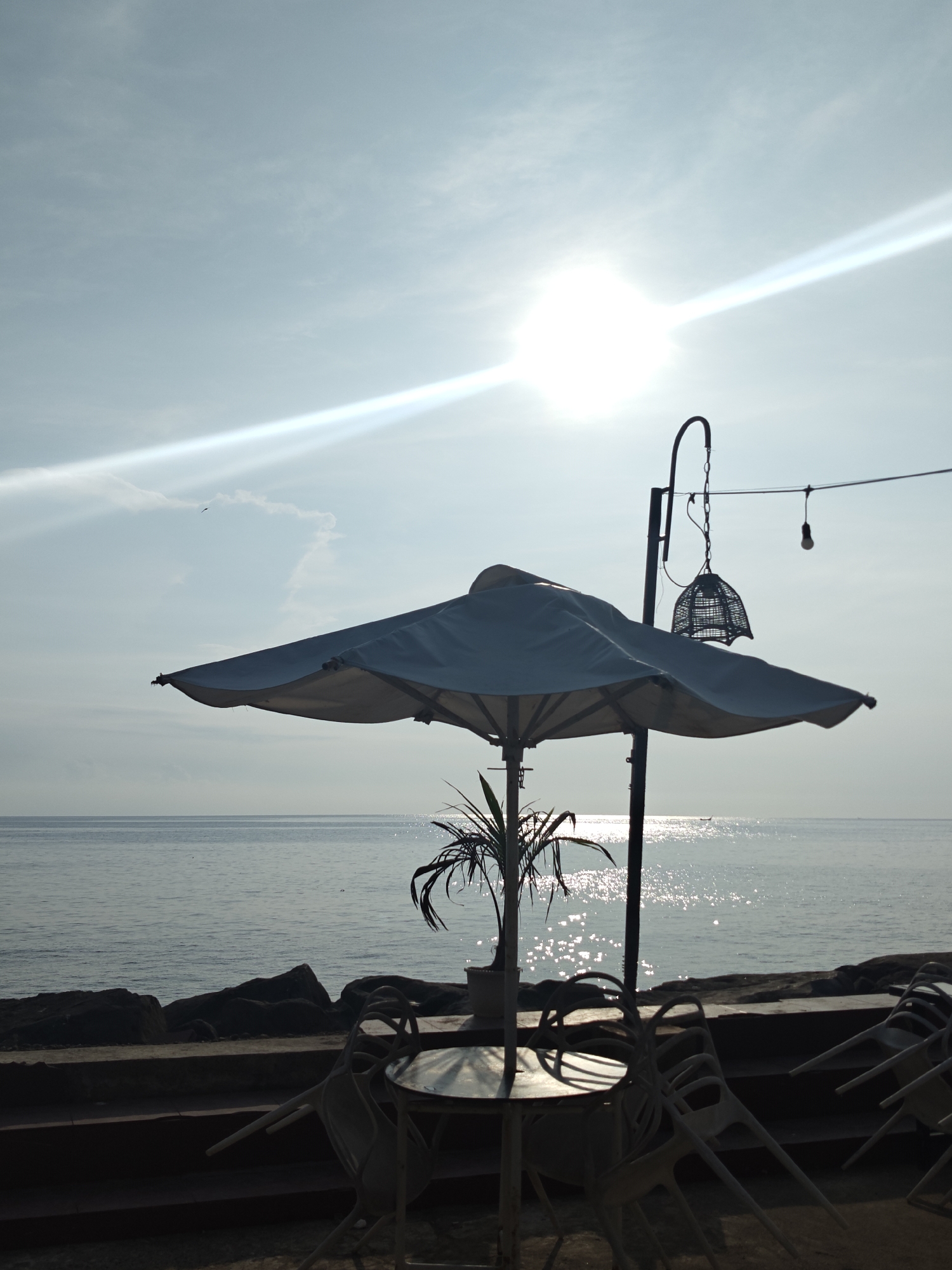 A seaside scene in Lhokseumawe, Indonesia, features a white parasol shading a table and chairs. The scene is cast in silhouette against the bright sun reflecting off the sea. A small potted plant sits on the table. A decorative light fixture hangs nearby. The background includes rocks lining the shore and the sea stretching to the horizon.