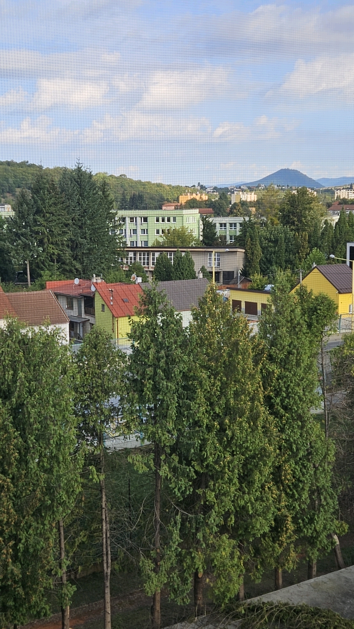 This is a medium-shot, vertical photograph of a cityscape with residential buildings and trees under a partly cloudy sky. The foreground is dominated by several tall, slender evergreen trees with dense green foliage. Behind the trees, a row of houses with red and brown tile roofs is visible, some with visible chimneys and satellite dishes. One house in this row is painted bright yellow. Further back, a long, light green building with multiple windows is situated, followed by other buildings of varying sizes and colors, including a lighter yellow structure with a dark roof and a building with a grey facade. In the distance, a rounded, tree-covered hill or mountain can be seen, with other hills and scattered buildings on the horizon. The sky is a pale blue with scattered white clouds. The photograph appears to have been taken from a slightly elevated position, possibly a window, as indicated by a faint grid pattern superimposed on the image. The scene depicts a residential area in a town or city, with a mix of older and newer architecture nestled among abundant greenery. The lighting suggests it is daytime, likely late afternoon or early morning given the soft light. The location is Prešov, Slovakia.