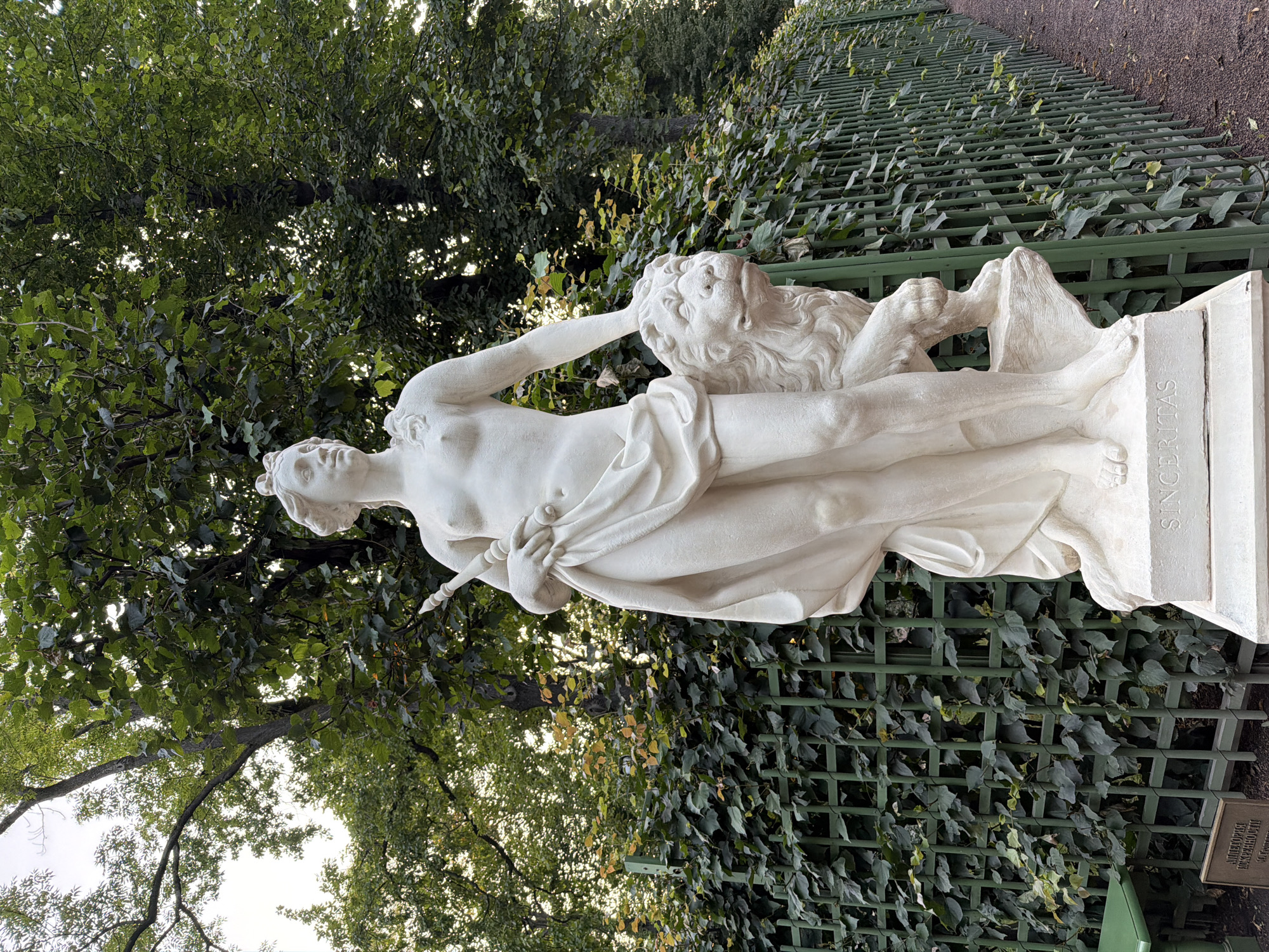 The image displays a full-body white marble statue of a female figure with a lion at her side, set against a backdrop of lush green trees and a green trellis fence covered in ivy. The statue stands on a rectangular pedestal inscribed with the word "SINCERITAS". To the left of the statue, a small green plaque is visible on the ground. The statue depicts a nude woman with her left arm resting on the lion's head and her right hand holding a scepter-like object. She is draped in flowing fabric. The environment suggests an outdoor setting, likely a park or garden. The sign on the ground likely provides information about the sculpture. The statue is located in the Summer Garden in Saint Petersburg, Russia.