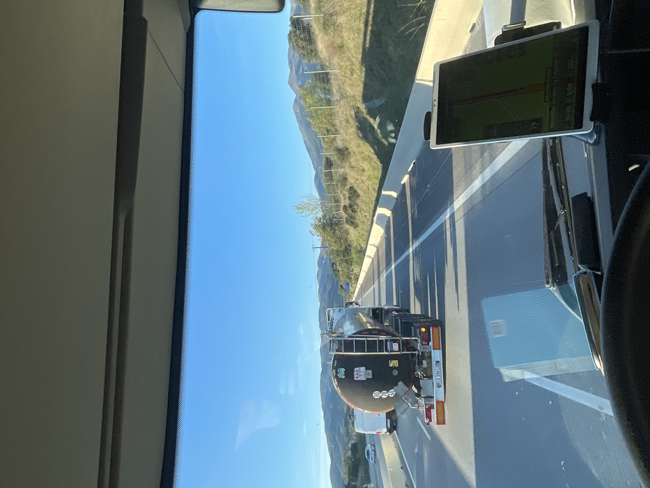 The image depicts a forward-facing view from the interior of a vehicle, likely a truck, traveling on a multi-lane highway near Maureillas-las-Illas, France. A clear blue sky is visible above. In the foreground, a portion of the vehicle's dashboard and steering wheel is present, with a white tablet device mounted vertically on the right side of the dashboard, displaying a green-hued navigation map. Directly ahead, a black tanker truck with a silver cylindrical tank is positioned in the right lane, featuring an "80" speed limit sticker and hazard placards on its rear. Reflections of the tanker truck and the sky are visible on the vehicle's windshield. A white van and another light-colored vehicle are visible in the left lane further down the highway. The roadway is marked with white solid and dashed lines. To the right of the highway, a light-colored concrete barrier separates the road from a grassy embankment with scattered trees and a wire fence. Mountains or hills are visible on the horizon. Long shadows are cast across the highway from the barrier and vehicles, indicating a low sun angle.