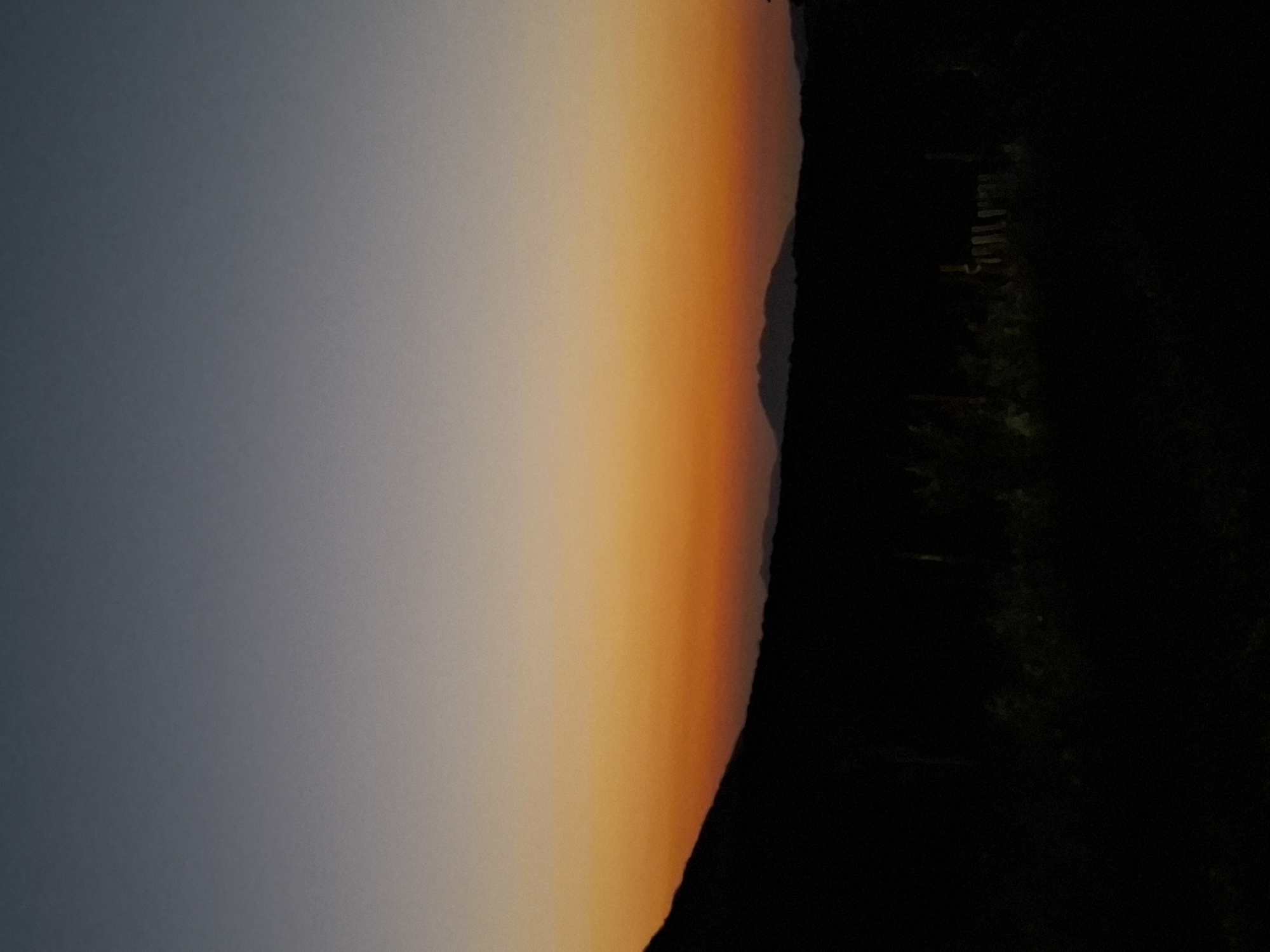 This is a photograph taken at dusk in Sel'mentauzen, Russia. The image depicts a landscape with a gradient sky transitioning from dark blue at the top to vibrant orange near the horizon. Silhouetted against the sky are rolling hills and mountains in the distance. In the foreground, a dark, dense line of trees and vegetation obscures the ground. A faint, dark silhouette of a fence is also visible within the foreground foliage, suggesting a rural or natural setting. The overall impression is one of a serene and natural environment at the end of the day.