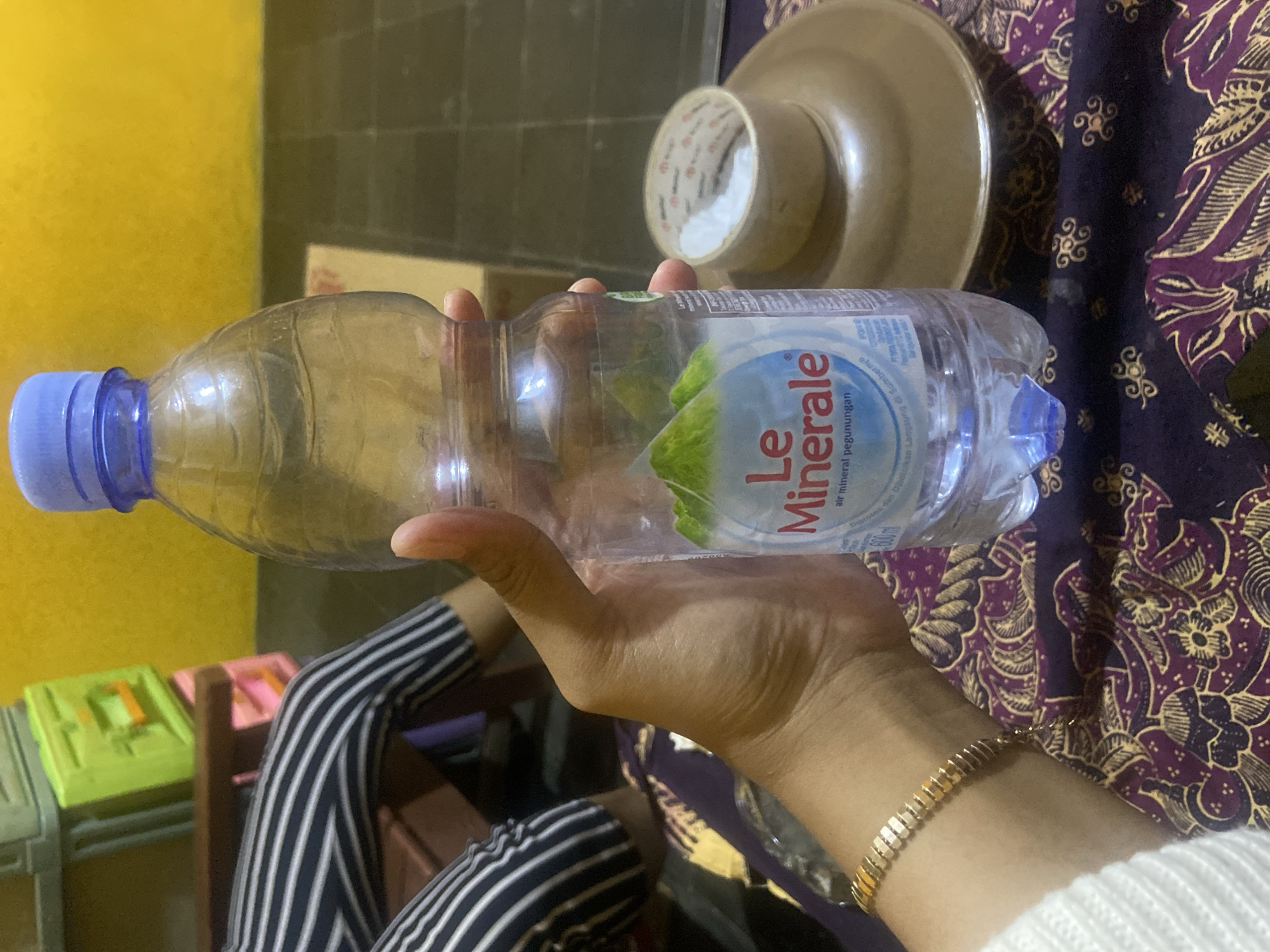 A person's hand holds a partially empty plastic bottle of Le Minerale bottled water, its blue cap visible. The hand wears a thin gold bracelet. The background is a yellow wall and black tiled floor. To the left, a person's legs wearing black and white striped pants are visible, seated near brightly colored plastic storage containers. To the right, a brown saucer holds a roll of white tape. The scene is indoors, potentially in a home or a small shop, based on the objects and furniture. The provided location information, Kecamatan Pati, Indonesia, suggests this is an everyday setting within a residential or commercial area of that region.