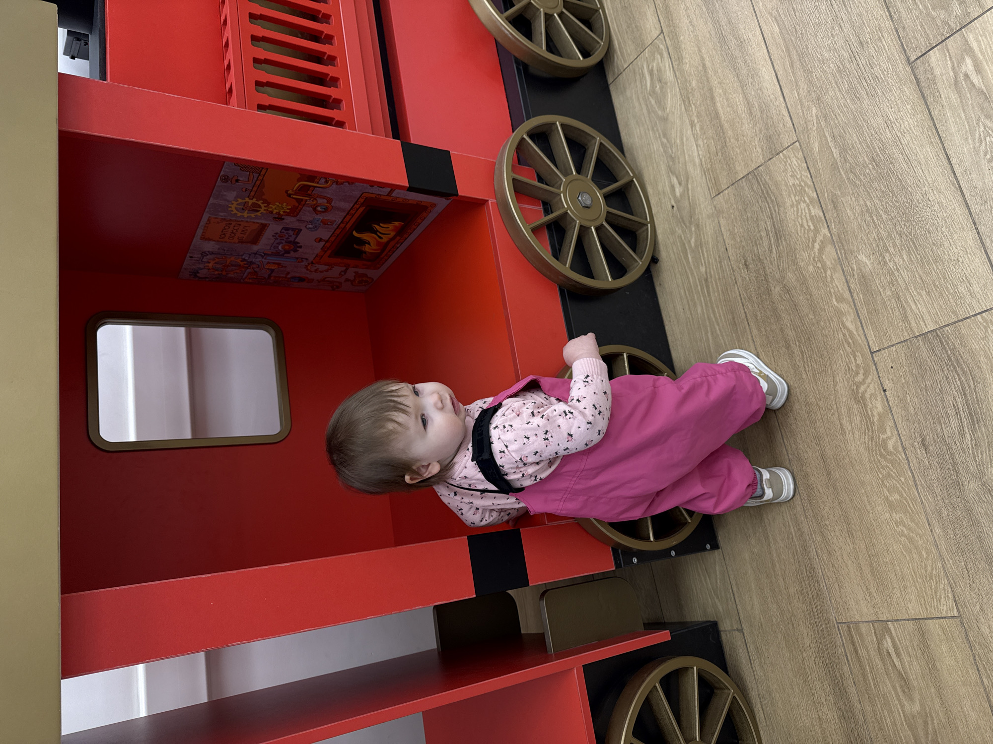 A toddler is playing on a wooden train toy. The toddler is wearing pink overalls and a floral shirt. The train is red with gold wheels. The flooring is a light brown wood. This is an interior scene. There is no indication of this being located in Moscow, Russia.