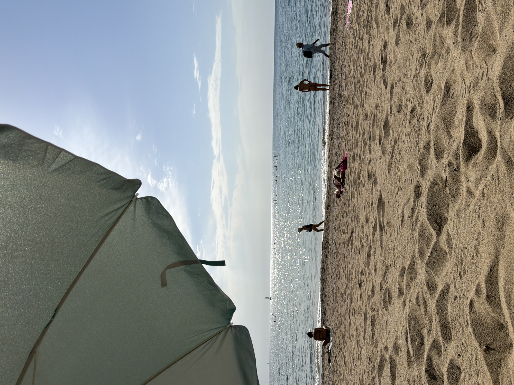 The image depicts a sunny day at a beach in Barcelona, Spain, with the foreground dominated by sand dunes and shadows cast by a large, light green beach umbrella in the upper left corner. The midground shows the shoreline with several people. One person is sitting cross-legged on a towel near the water's edge, facing the sea. Further down the beach, a person in a bikini stands with hands on hips, and another person in light-colored clothing walks away from the camera carrying a backpack. In the background, the Mediterranean Sea sparkles under the bright sun, with several figures visible in the water, some appearing to be paddleboarding. Sailboats are also visible on the horizon. The sky is a clear blue with a few wispy clouds. The overall scene conveys a relaxed atmosphere of a summer day at the beach.