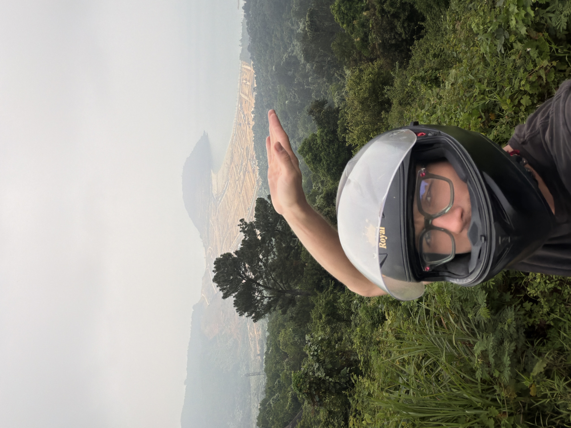 A person wearing a motorcycle helmet and glasses is positioned in the foreground, their right hand raised. In the background, a hazy view reveals a coastline and a sprawling development area, with trees and vegetation partially obscuring the scene. Based on the landscape features, the location is potentially in or around Đà Nẵng, Vietnam. The environment suggests a vantage point along a road or hillside overlooking the coastal region.