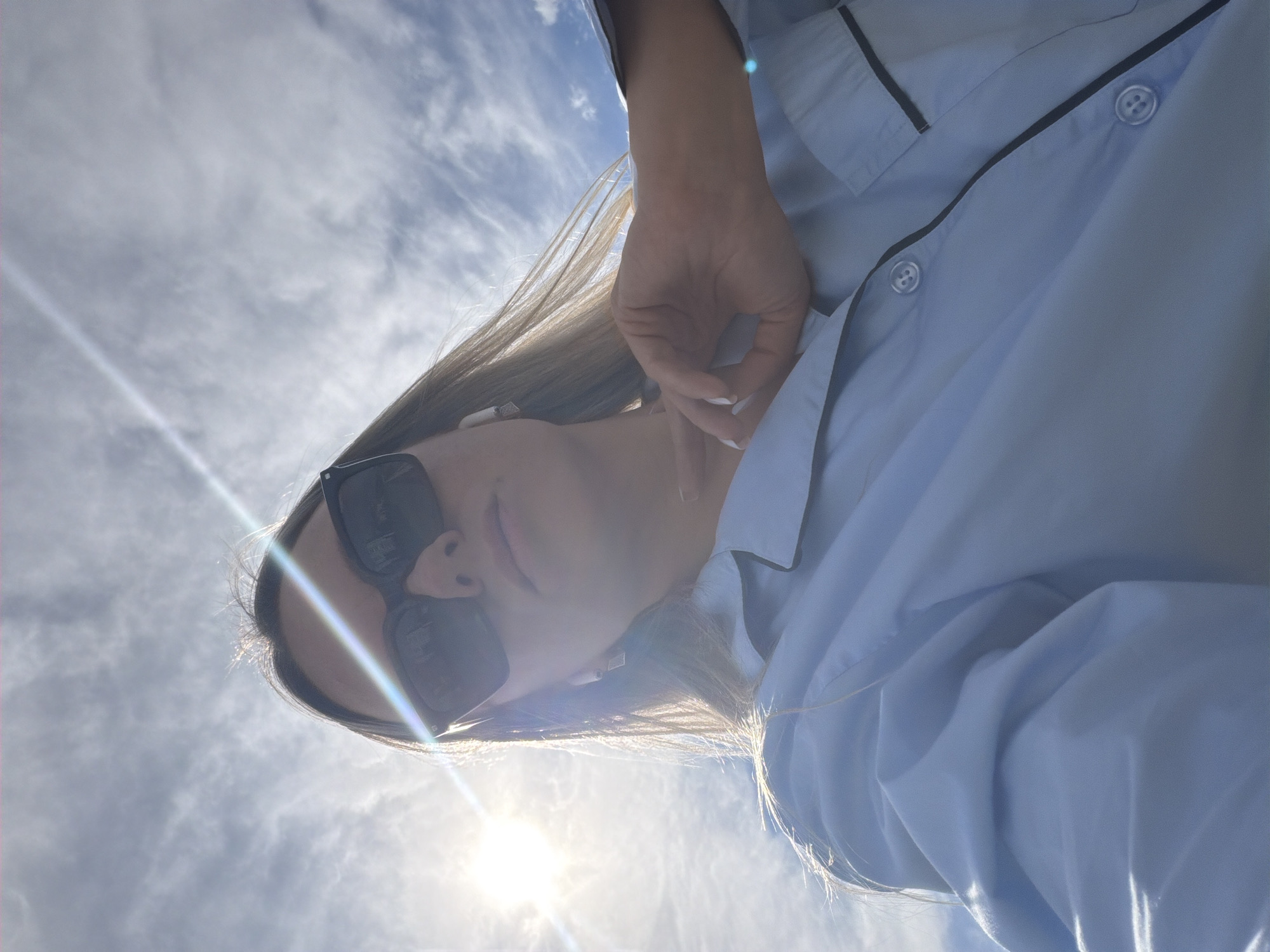 The original image, taken from a low angle, captures a person against a bright, cloudy sky in Votkinsk, Russia. The individual, likely female, is positioned diagonally across the frame, looking downwards towards the camera, suggesting a selfie or a shot taken from below.
The subject has long, light brown hair, some strands of which are illuminated by the sun. She is wearing dark, rectangular sunglasses that obscure most of her eyes. Her lips are slightly parted. She is dressed in a light blue collared shirt with thin, dark piping along the collar and front placket. Her left hand, with nails painted white, is raised near her face, possibly resting lightly or adjusting her hair.
The photograph depicts an outdoor setting during the daytime. The sun is intensely bright, located towards the lower-left of the frame, creating significant lens flares that streak across the image and highlight the person's hair. The sky is scattered with wispy white clouds, indicating clear weather. The overall impression is casual and bright. No specific location cues for Votkinsk, Russia, other than the sky, are visible. There is no visible text in the image.