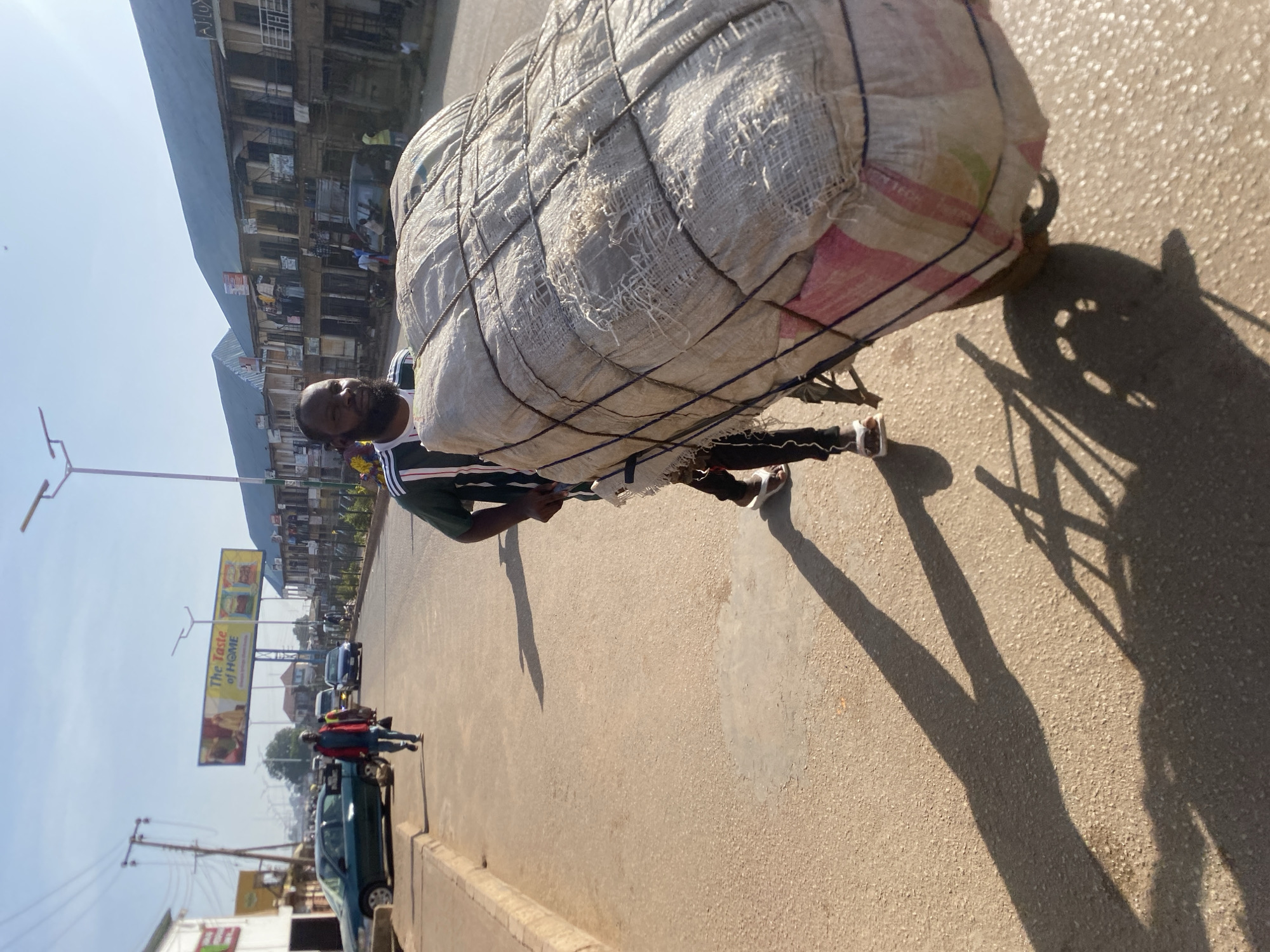 In the city of Bukuru, Nigeria, a man is captured in a medium shot, walking on a paved road and pushing a large, bundled load on a wheelbarrow. The load is wrapped in a rough, light-colored fabric and secured with dark ropes. The man is wearing a dark green and white striped t-shirt and dark pants, with white sandals. His face is visible, and he appears to be looking forward. Behind him, the road stretches into the distance, lined with buildings that appear to be shops or residences with multiple stories and balconies. A billboard with the text "The Taste of Home" is visible in the background. Several cars and other pedestrians are also present on the road, indicating moderate activity. The scene is illuminated by bright sunlight, casting long shadows on the ground. The overall environment suggests a commercial or residential street in an urban setting.