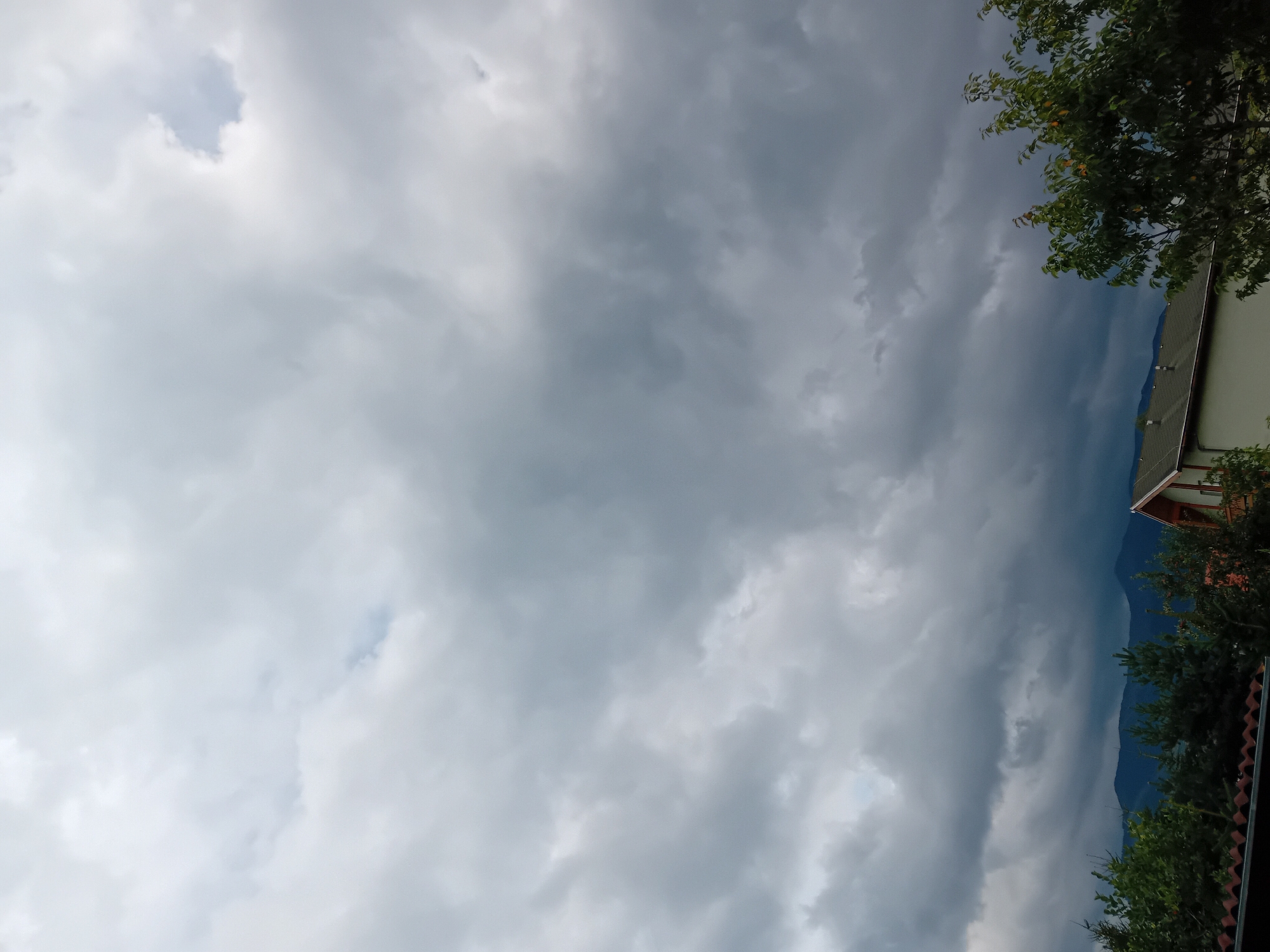 The image depicts a sky filled with heavy, dark clouds, suggesting an impending storm or overcast weather. In the foreground, there are trees with green foliage and a partial view of rooftops, including a house with a white wall and a brown roof. In the distance, a range of dark blue mountains is visible. The scene appears to be set in a rural or suburban area, consistent with the location of Zuberec, Slovakia, which is known for its mountainous landscape and traditional architecture. There are no people or animals visible in the frame. The overall impression is one of a dramatic, cloudy sky over a mountainous region.