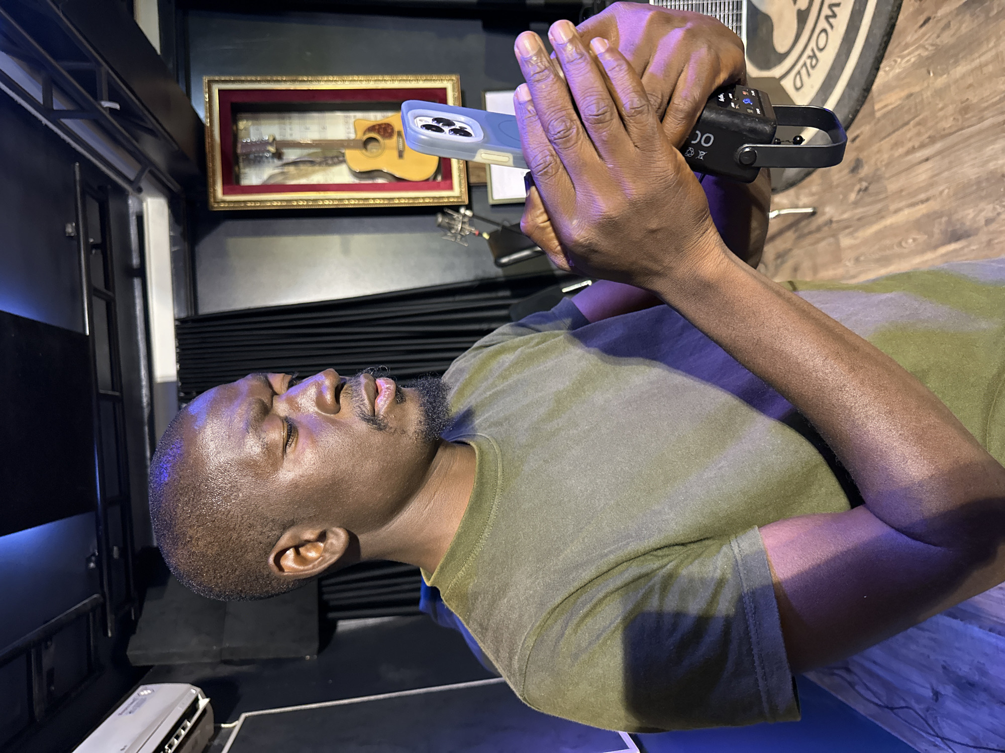 A man of African descent is shown in a medium close-up, holding a mobile phone in his hands. He is wearing a plain olive green t-shirt. The background appears to be a studio setting with dark curtains and a framed acoustic guitar displayed on the wall. There is also an air conditioning unit visible on the left side of the image. The man's gaze is directed downwards at the phone, suggesting he is interacting with it. The lighting in the scene is dim, with some colored light casting shadows on his arm and body. The overall impression is of a recording or creative space. The image does not contain specific elements to identify a location in Barakin Lerawi, Nigeria.