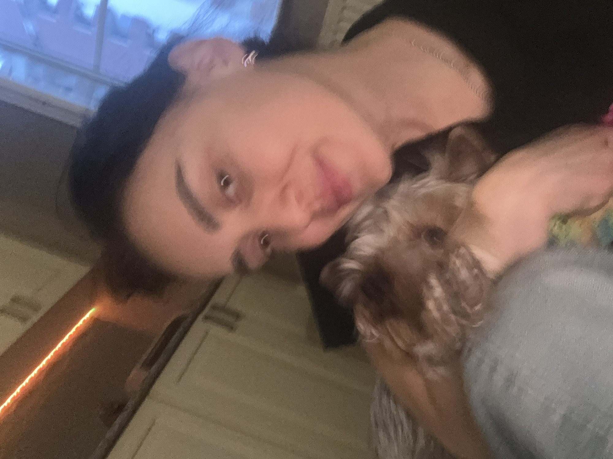 A woman with dark hair and pierced nose and ears is holding a small dog. They are indoors, possibly in a kitchen, as indicated by cabinets and LED strip lights in the background. It is not possible to verify the location in the city of Tbilisi, Georgia, from the given image.