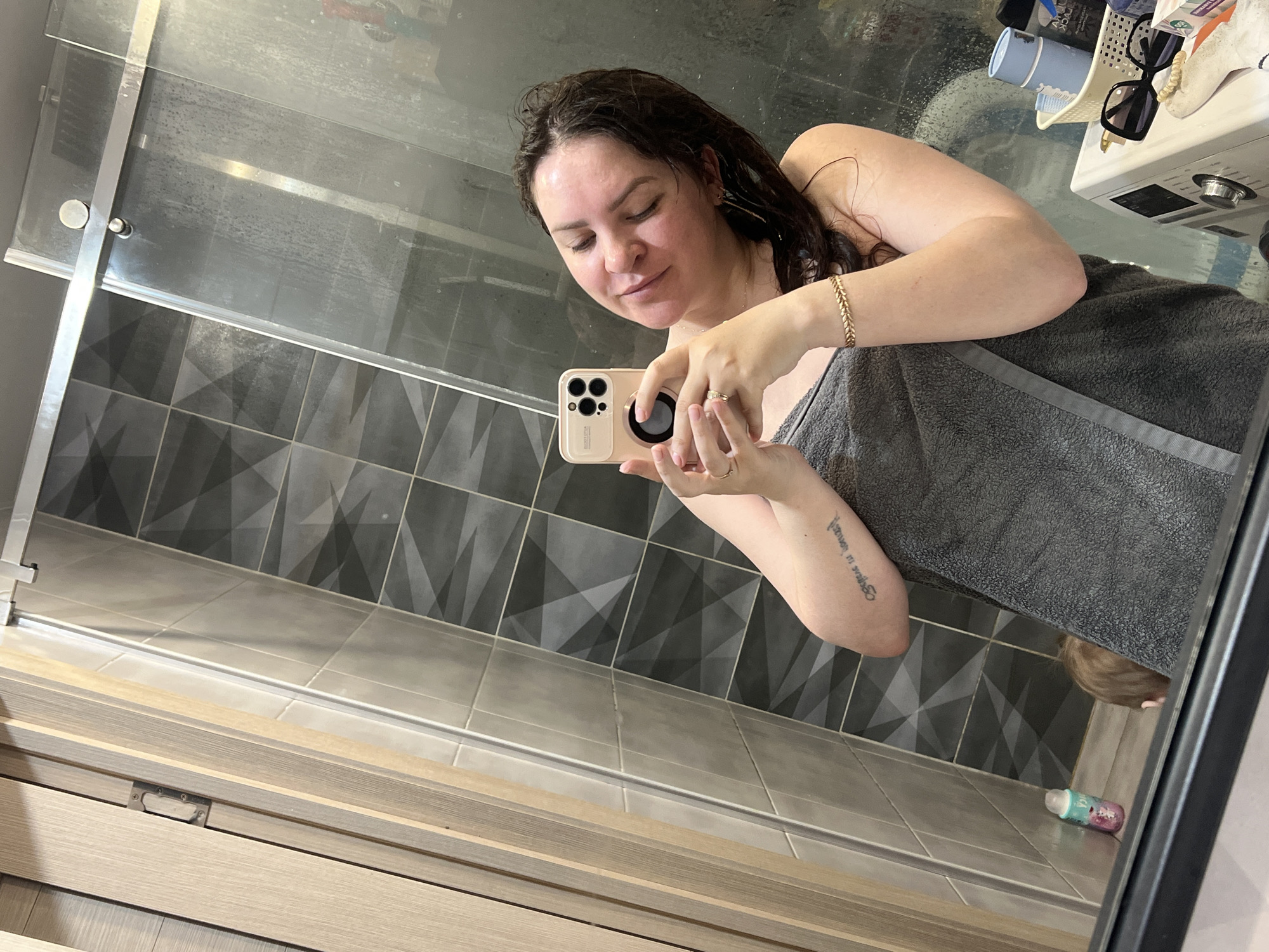 A woman stands in a bathroom, wrapped in a grey towel, holding a smartphone to take a selfie in the bathroom mirror. Her hair appears wet. The bathroom features grey patterned tiles on the wall and lighter-colored tiles on the floor. A shower door is visible, as is a cabinet or counter top with various items, including sunglasses and toiletries. On the floor below, a child's head and a small container are visible. The location in the city of Noviy, Russia, cannot be determined from this image.