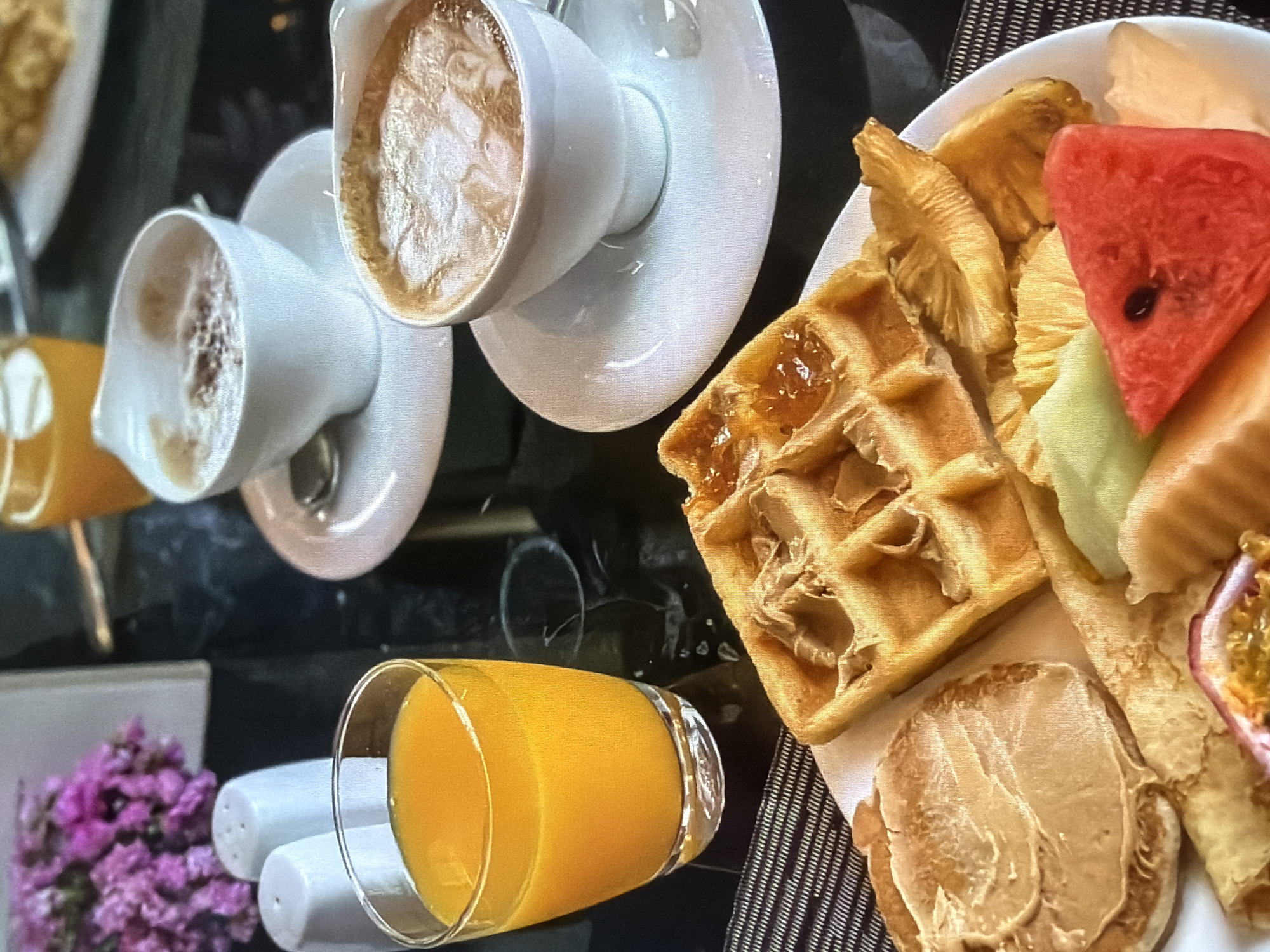 The image captures a vibrant and varied breakfast or brunch spread, presented on a dark, reflective table surface with a textured, dark gray placemat. The scene is likely indoors, possibly in a hotel, cafe, or restaurant setting.
Prominently featured are several food and drink items. Two white ceramic coffee cups with matching saucers are filled with coffee, displaying intricate foam art on their surfaces. A clear glass holds bright orange juice. A white plate showcases a selection of breakfast foods: a golden-brown waffle, partially covered with a glistening amber-colored jam or syrup. Alongside the waffle, there is an assortment of fresh fruit including pieces of pineapple, a striking red triangular slice of watermelon with a visible seed, lighter-colored melon pieces (possibly honeydew or cantaloupe), and a half passion fruit revealing its characteristic seedy pulp. Also on the plate are a folded thin pancake or crêpe, and a round, toasted item (resembling a bagel half or a thick pancake) generously spread with what appears to be peanut butter or a similar nut spread.
Other objects in the frame include two white salt and pepper shakers and a small decorative arrangement of purple flowers. The image is taken from a slightly overhead perspective and appears tilted, adding to the dynamic composition. There are no people visible, and no visible text in the image. The time of day suggested by the meal items is morning or early afternoon. The context provided indicates this scene is in Lovech, Bulgaria.
