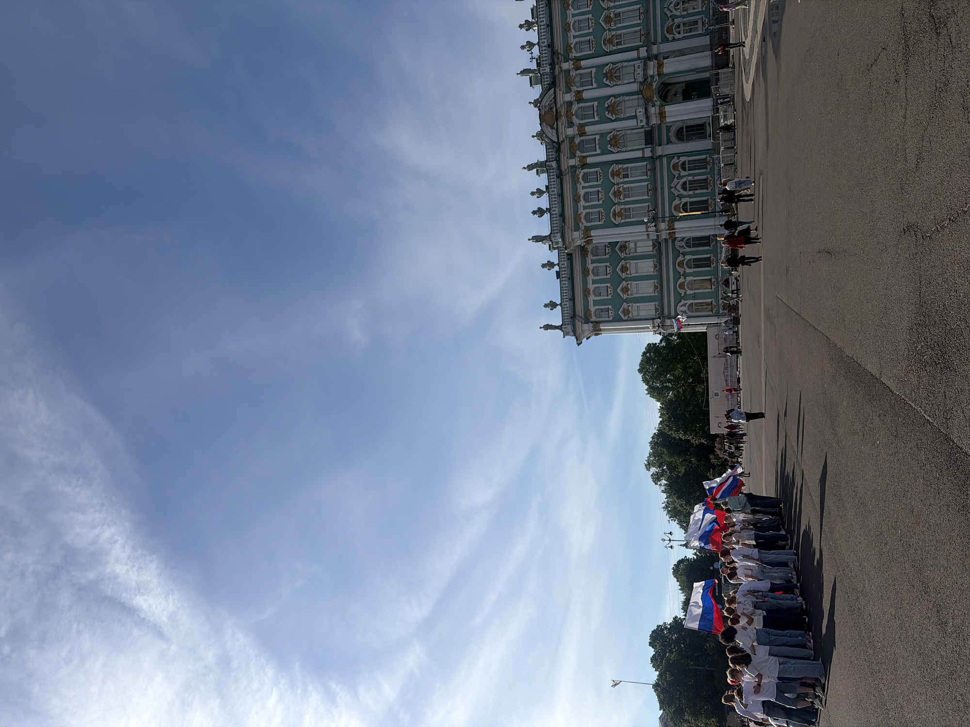 A group of people, likely tourists or participants in an event, stand in a line facing a large, ornate building. Several of the people are holding Russian flags. The building, which appears to be historical, is green and white with many windows and decorative elements. The scene is outdoors, with a paved area in the foreground and trees in the background. The sky is blue with wispy clouds. The overall impression is a sunny day at a notable landmark.