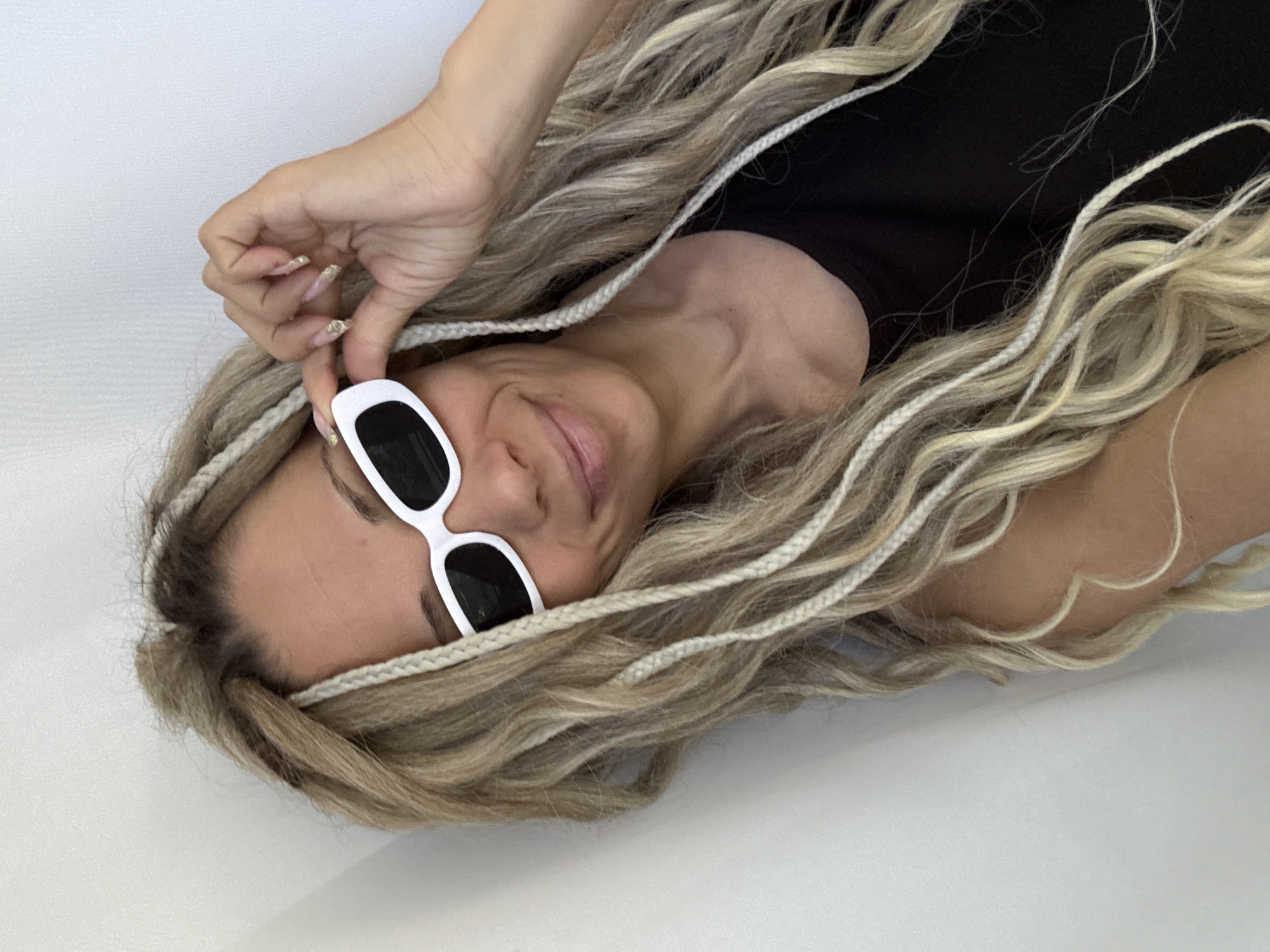 A woman is lying down, angled towards the upper left of the frame. She is wearing a black top and white-framed sunglasses. Her blonde hair is styled with small braids. Her right hand is near her face, adjusting the glasses. The background is a plain white surface. There is no indication of the location being in the city of Rusanovo, Russia.