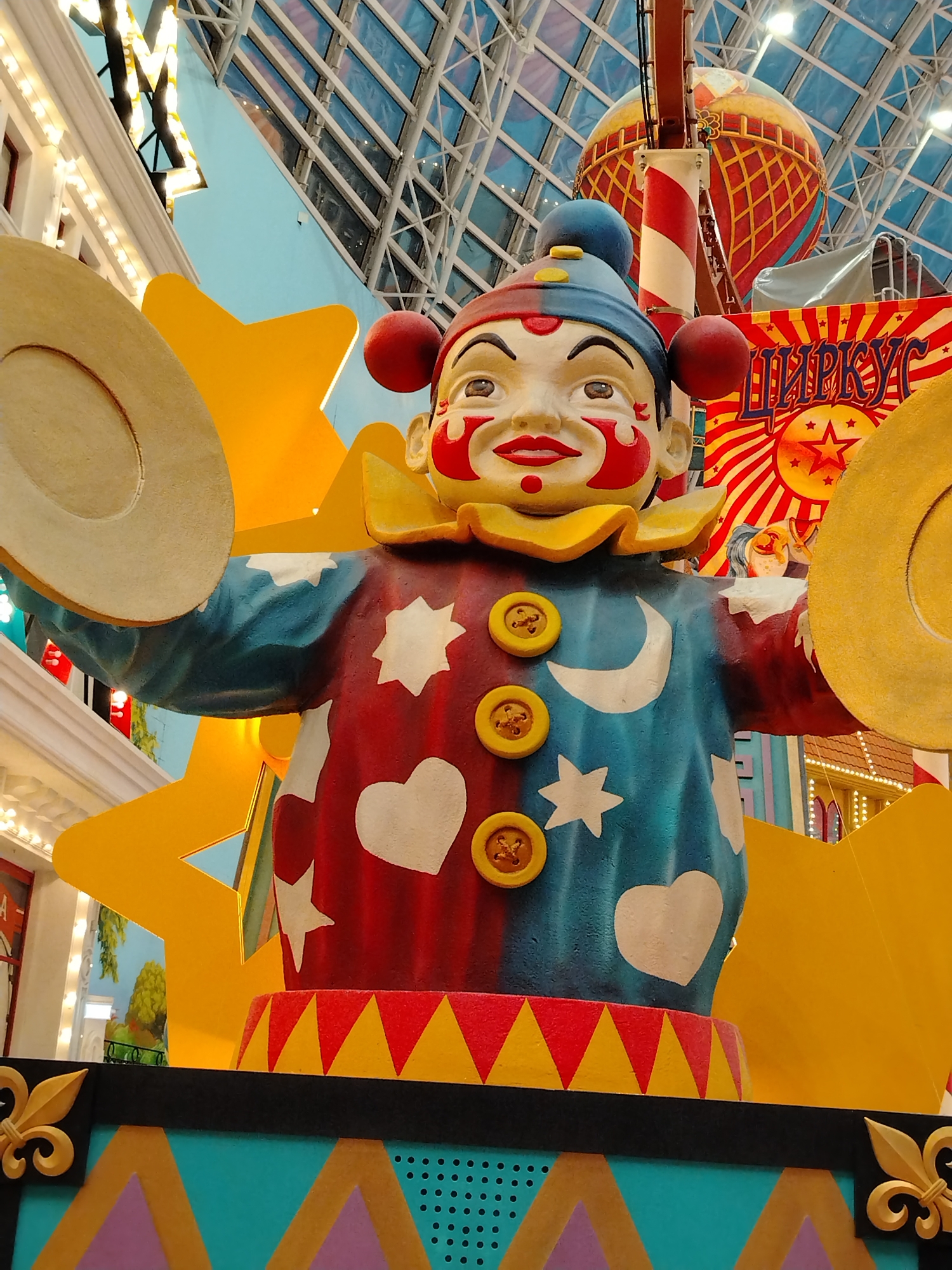 The image shows a life-size clown figure in a brightly colored, indoor setting that appears to be a circus or amusement park. The clown is dressed in a red and blue outfit adorned with white stars and hearts. It has large red cheeks, a smiling mouth, and is holding two large golden cymbals. The clown is positioned in front of a large yellow star decoration. In the background, there is a striped pole and a large hot air balloon-like structure, along with a banner with Cyrillic lettering that reads "ЦИРКУС" (circus). The ceiling is a geodesic dome made of glass and metal. The overall impression is of a festive and theatrical environment.