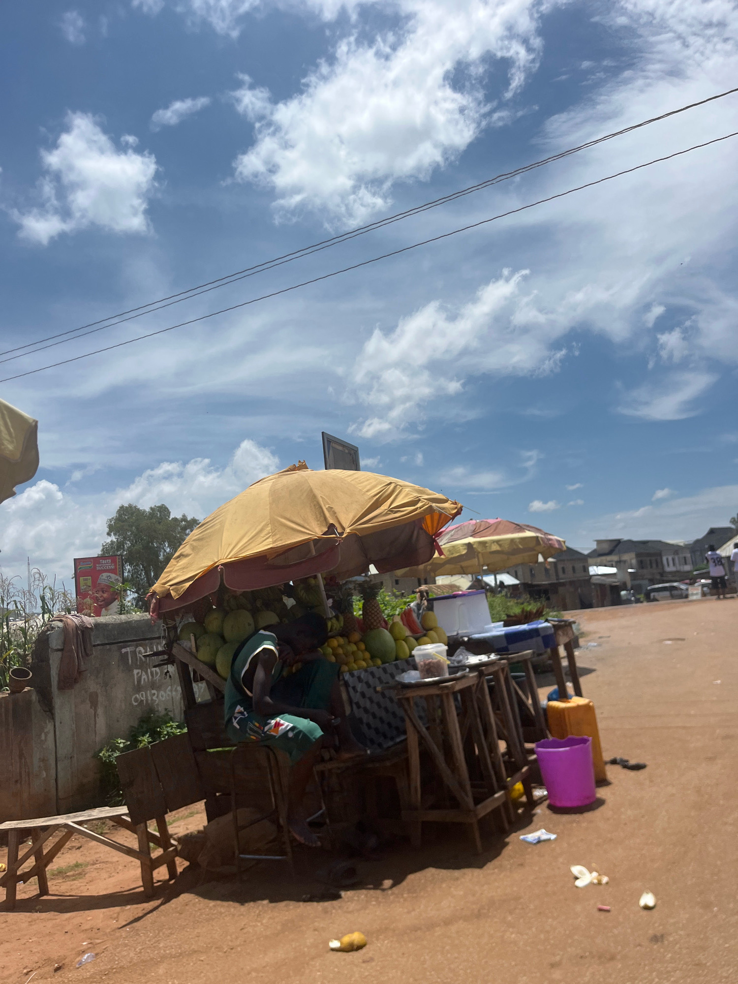 The image captures a roadside fruit stand in Rafin Par, Nigeria, under a partly cloudy sky. A vendor, a young man wearing a green and white sleeveless top and green shorts, is seated on a wooden stool behind a table laden with a variety of fruits, including pineapples, coconuts, oranges, and other produce. Two large umbrellas, one yellow and one pinkish-orange, provide shade for the stand and its contents. A wooden bench is visible to the left of the stand, and further left, a concrete structure has writing on it. In the background, houses and buildings are visible, and power lines crisscross the sky. The ground is a reddish-brown dirt path, littered with some debris. A purple bucket and a yellow container are near the stand.