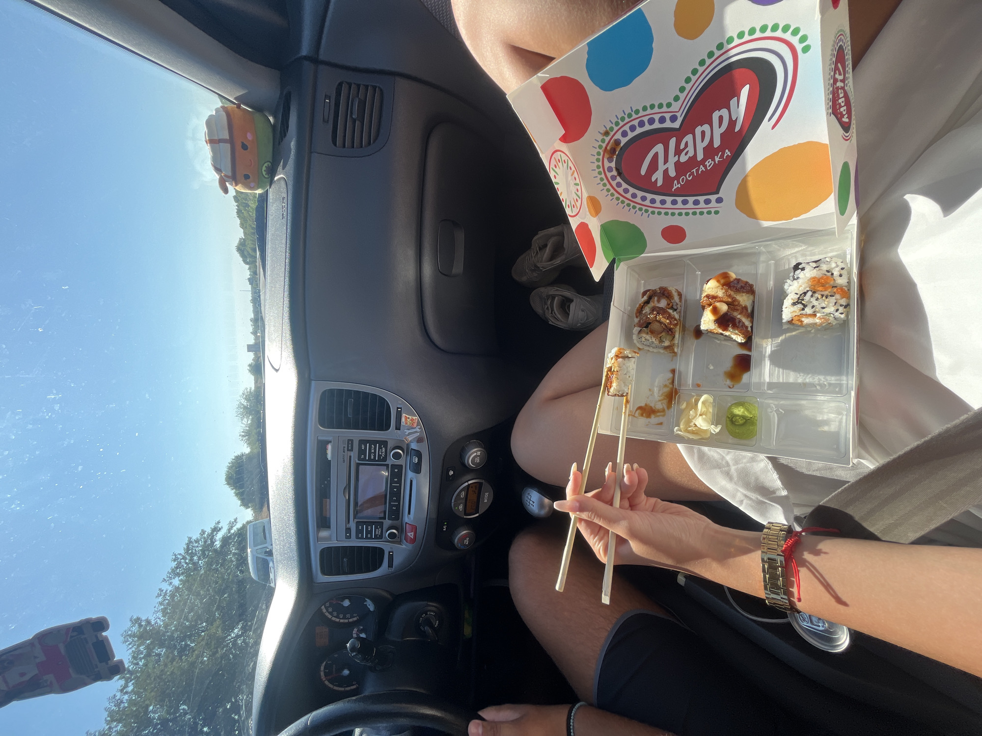 The image shows the interior of a car on a sunny day. Two people are seated in the front, with one person's hands holding chopsticks over a container of sushi. The sushi container is a white cardboard box with colorful polka dots and a heart-shaped logo that says "Happy Доставка" (Happy Delivery). The sushi inside appears to be several pieces, some with a glaze and others topped with sesame seeds. There are also small compartments with what looks like pickled ginger and wasabi. The car's dashboard is visible, with the steering wheel, radio, and air vents. Outside the car, trees and a clear blue sky are visible. One person's legs are visible, wearing dark shorts, and the other person's legs are mostly obscured by a white garment. A pair of gray sneakers are placed on the floor of the car. A small plush toy resembling a dessert is visible on the dashboard. The scene does not provide any specific visual cues that would indicate the location as Cherna Gora, Bulgaria.