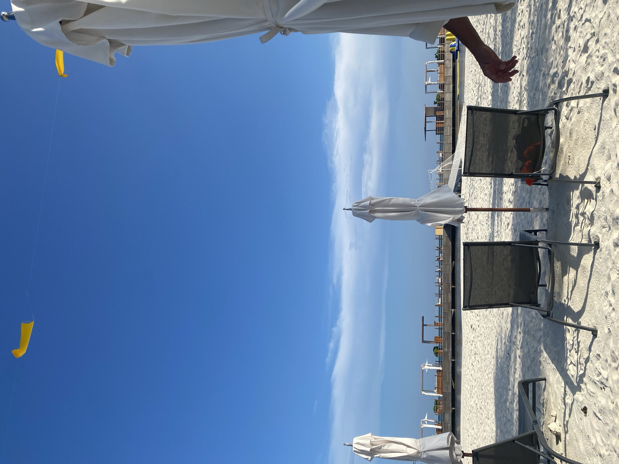 The image shows a beach scene in Marina Maroma, Mexico. Three closed, white beach umbrellas are present along the sand, with accompanying dark-colored beach chairs arranged in rows. The sky is predominantly clear blue with streaks of white clouds. A hand is partially visible at the upper right edge of the image. A yellow object is dangling in the sky. The beach appears to be lined with structures and vegetation in the background.