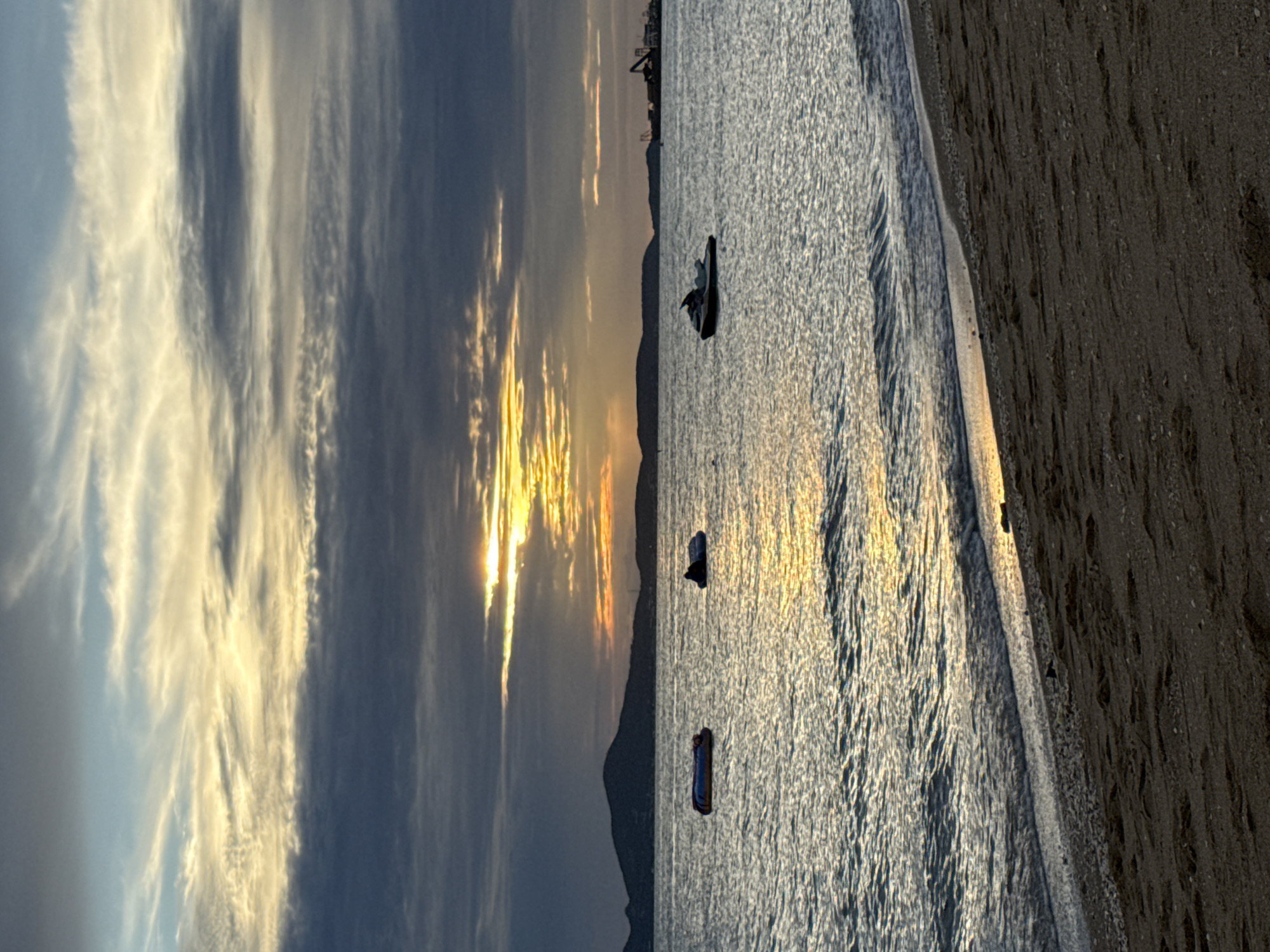 This is a vertical, eye-level shot of a tranquil beach scene in Pattaya City, Thailand, during what appears to be sunset. The sky is a dramatic canvas of dark gray clouds interspersed with streaks of golden light, hinting at the sun’s descent behind them. The sea is calm, reflecting the sky's colors with shimmering ripples.
In the water, there are three distinct objects. To the left, a blue and orange inflatable tube floats, empty. To its right, a dark, round object, possibly another inflatable or a person on a buoyant object, is partially submerged. Further to the right, a white and black jet ski is stationary on the water. In the distance, a dark, silhouetted mountain range lines the horizon. Beyond the mountain, on the far right, industrial structures and cranes are visible, suggesting a port or industrial area.
The foreground of the image is dominated by a sandy beach, with small shells and pebbles scattered across its surface. The sand is dark and appears damp near the water's edge, where gentle waves are lapping. The overall impression is one of peacefulness, with the soft glow of the setting sun casting a serene atmosphere over the scene.