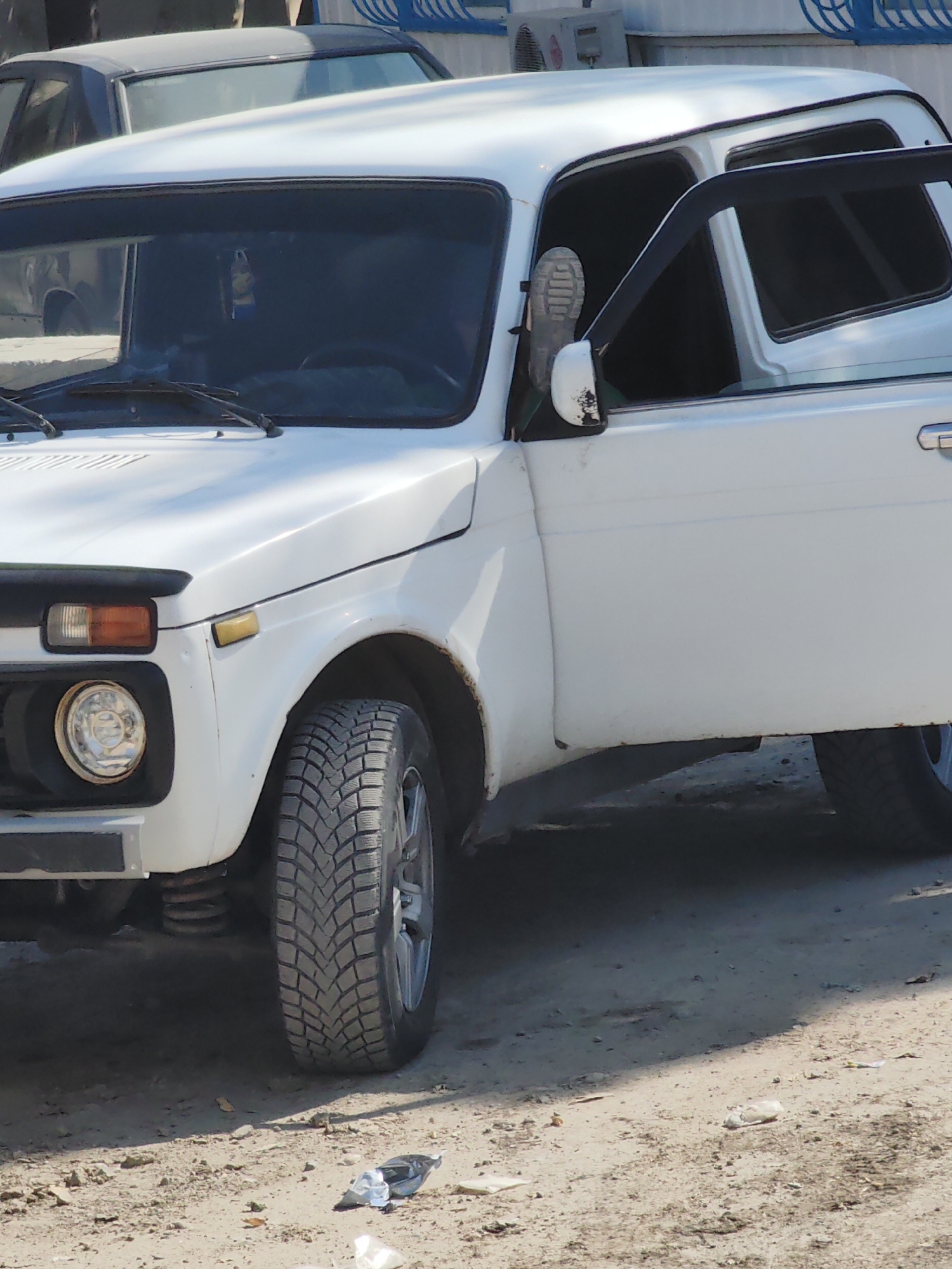 A white Lada Niva is parked on a dirt and gravel surface, with its driver's side door open. A person's foot is visible extending out of the open door. The car's headlights, turn signals, and tires are visible. In the background, another vehicle, a dark-colored sedan, is partially visible. The scene appears to be in an outdoor, urban environment.