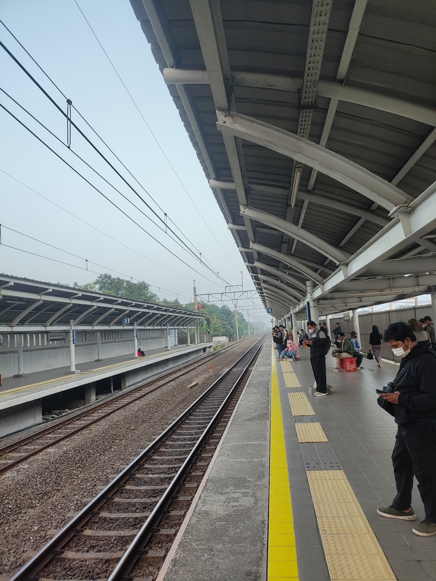 The image depicts a train station platform in Telagamurni, Indonesia. Several individuals are waiting on the platform, with many wearing face masks. The platform is covered by a gray metal roof supported by white pillars. Train tracks run alongside the platform, with gravel ballast beneath the rails. The sky is overcast and hazy. The platform features tactile paving for visually impaired passengers and a yellow safety line at the edge. One sign on the platform indicates "Mutiara Tiga Mulya". Several people are looking at their phones, while others are seated or standing.