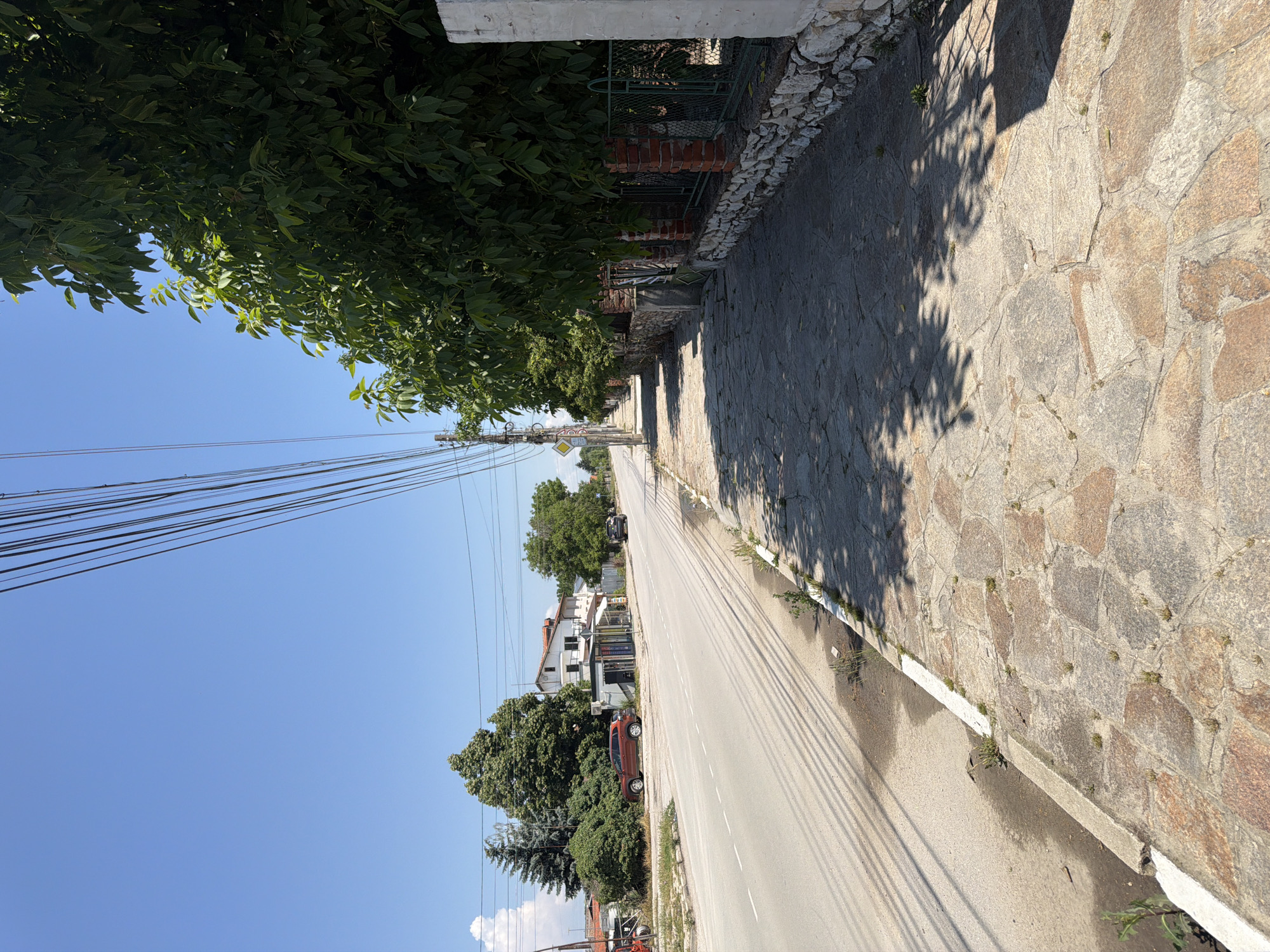 This is a photograph taken on a sunny day in Zlatitrap, Bulgaria, showing a paved sidewalk alongside a road. The sidewalk is constructed with irregularly shaped, flat stones of various earthy tones and has patches of small green weeds growing between them. The road is asphalt and has a white dashed line marking the lanes.
To the left of the road, there are some trees, buildings, and a red SUV. The buildings appear to be residential or commercial. Overhead, numerous electrical wires stretch across the sky, supported by poles.
To the right of the sidewalk, there is a low wall constructed with rough stones and brick pillars, topped with a green metal fence. Beyond the fence are trees with dense green foliage, casting shadows on the sidewalk. A white concrete pillar is visible on the far right of the image. The sky is a clear blue with a few wispy white clouds. The scene depicts a quiet street on a warm day, likely in a residential or semi-urban area.
