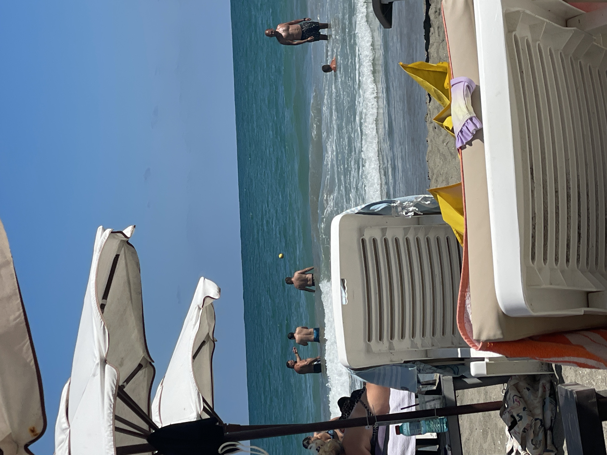 The image depicts a sunny day at a beach in Pomorie, Bulgaria. In the foreground, two white beach chairs are visible, one partially obscured. A patterned beach towel and a folded swimsuit in pastel colors rest on the back of the chair closest to the viewer. Beyond the chairs, several people are in the sea, with some wading and others swimming. Three men are in the water near the shore, with one appearing to point at something in the distance. Further out, a man stands in the water while a child swims nearby. The sea is a vibrant turquoise, with white waves crashing along the shoreline. The sky is clear blue. Beach umbrellas are visible in the upper left corner of the image, suggesting a resort or public beach area. The sand is a light brown color.