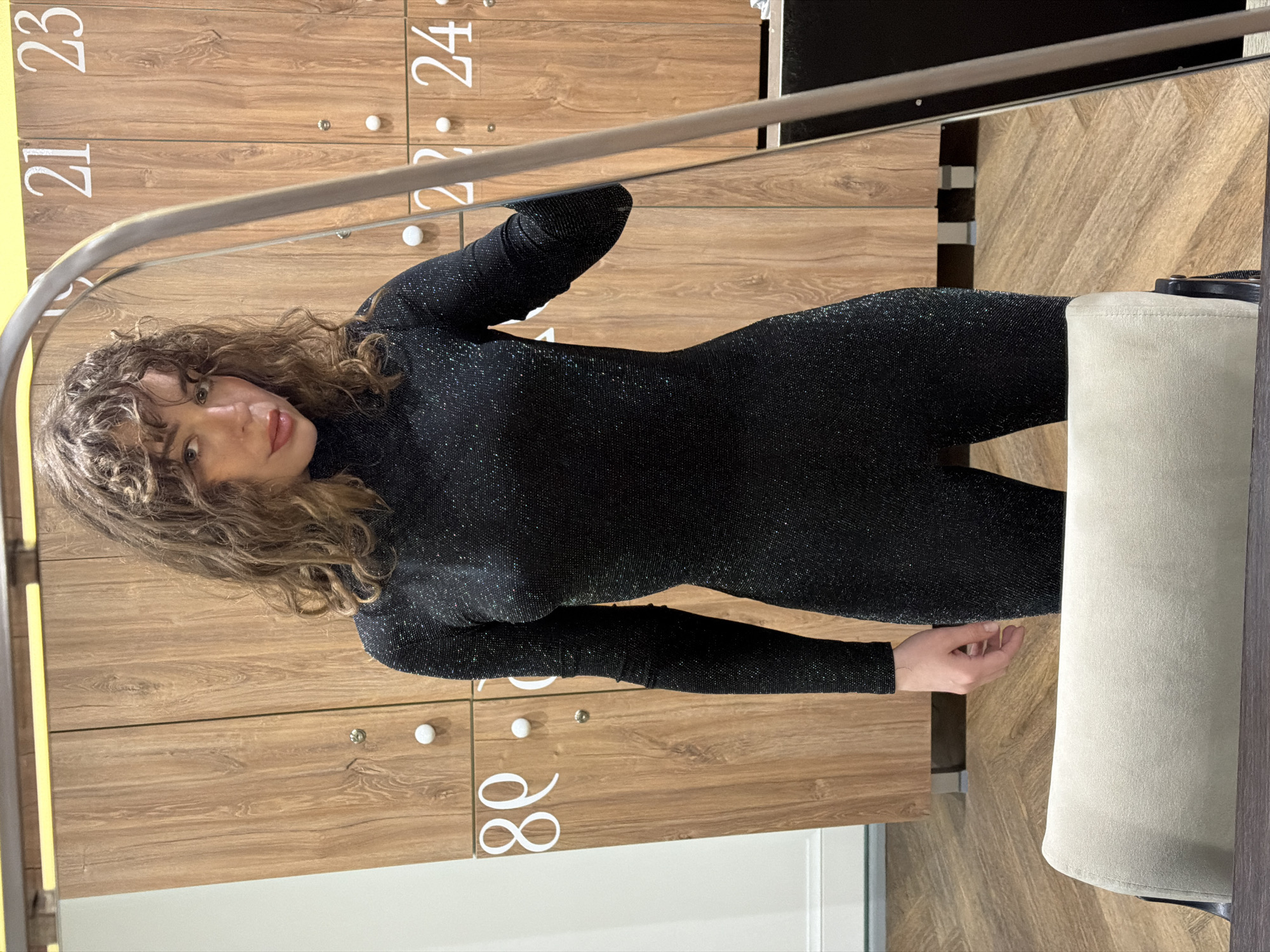 The image shows a woman with curly brown hair wearing a black, glittery bodysuit. She is standing in what appears to be a changing room or locker room, with wooden lockers labeled with numbers 21, 22, 23, 24 and 88. A partial view of a stool or chair is visible on the right. The location is Moscow, Russia, assuming the locker label is a reference to the city's area code (+7 495/499).