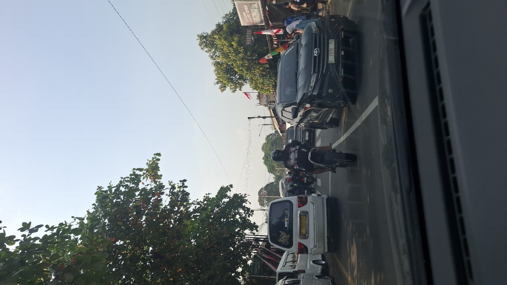 The image depicts a street scene in Cilaku, Indonesia, with moderate traffic. A white van is parked on the left, while a black SUV, a motorcycle, and other vehicles are on the road. A person wearing a helmet is riding the motorcycle. The sky is clear, and trees are visible on the left side of the image. A sign for "WARTEG SEDUNIA FAVORIT SEMUA" is visible on the right, indicating a local eatery. Indonesian flags are displayed on poles. The perspective is from inside a vehicle, looking forward.