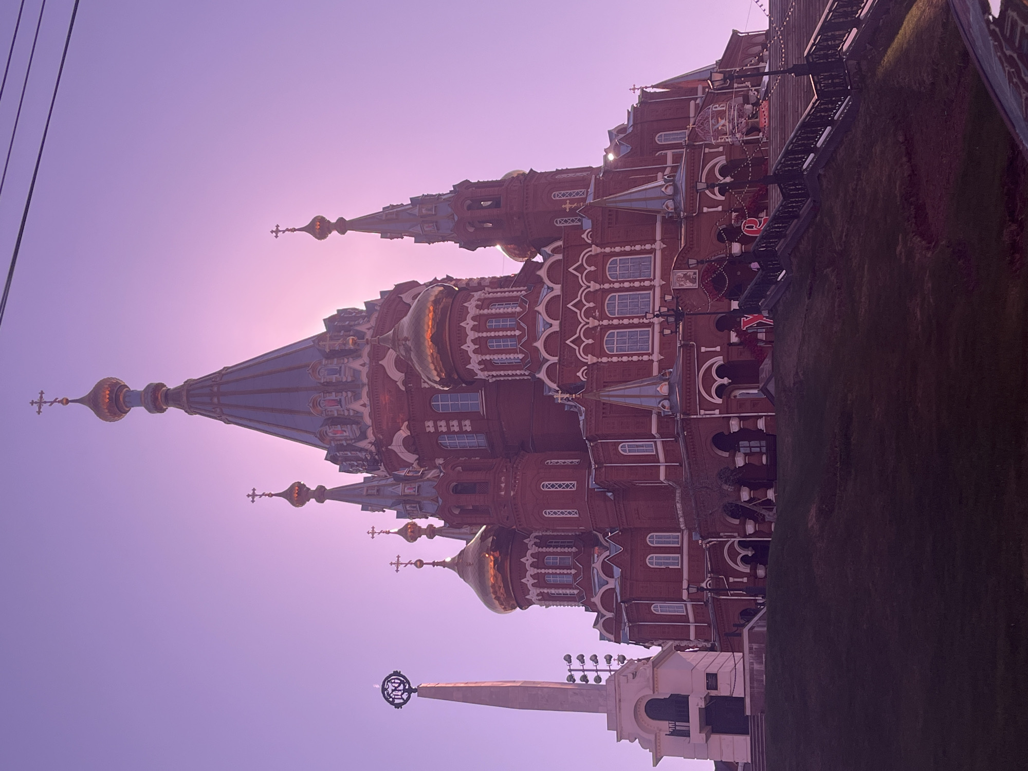 The image depicts the Cathedral of Alexander Nevsky in Izhevsk, Russia, at dusk or dawn. The cathedral is constructed of red brick, adorned with white decorative elements and multiple golden domes topped with crosses. The sky is a pale purple hue, suggesting the time of day. Power lines are visible in the upper left corner. To the right of the cathedral is a strip of green, likely grass, and a black fence. Another smaller building, white with a spire and emblem, is positioned to the lower right of the cathedral.