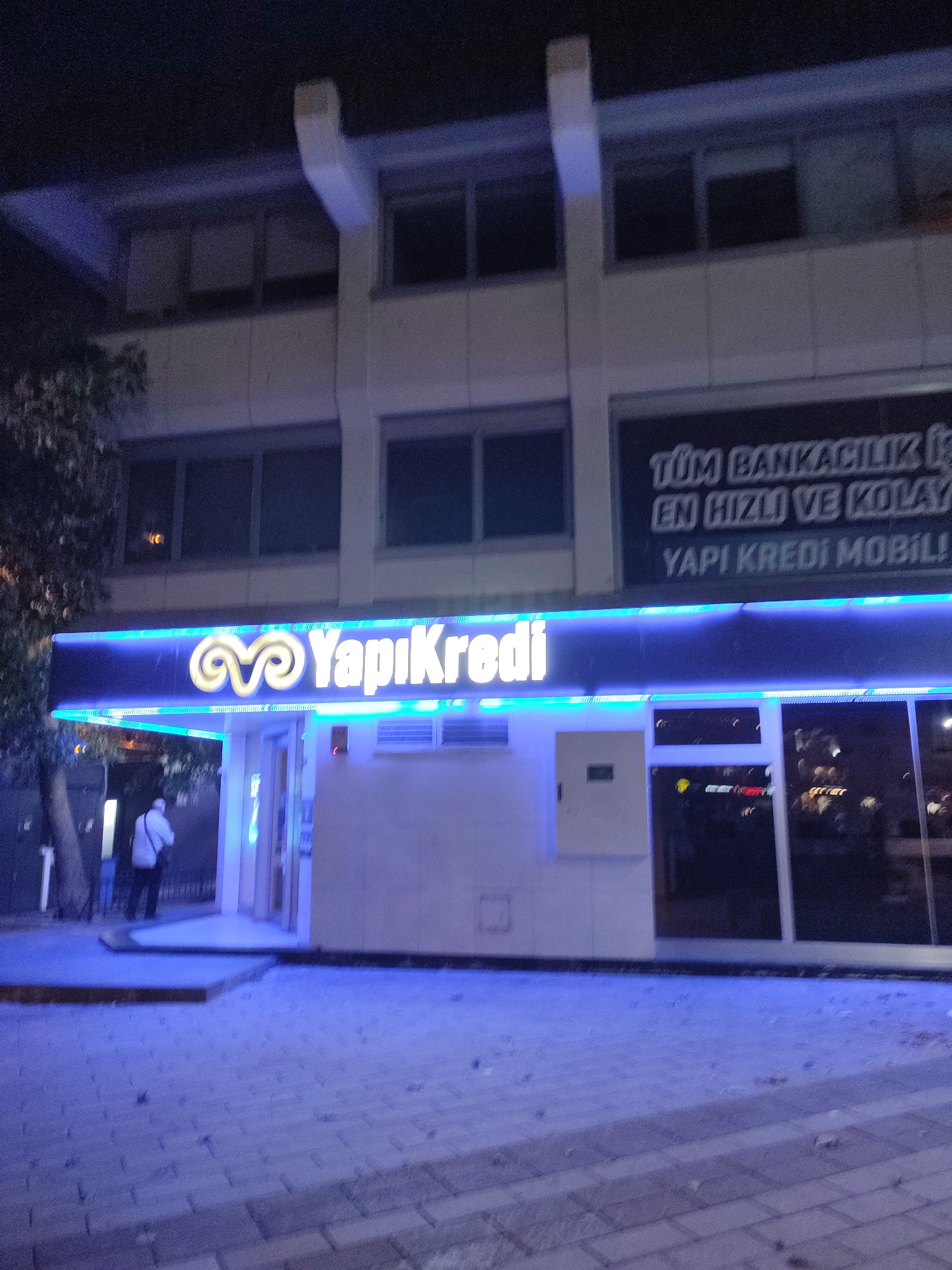 A nighttime exterior shot captures the facade of a Yapı Kredi bank branch in Üsküdar, Turkey. The building features a modern design with large windows and an illuminated blue neon sign that reads "Yapı Kredi". Above the entrance, another sign in white text displays "TÜM BANKACILIK İŞLEMLERİ EN HIZLI VE KOLAY YAPI KREDİ MOBİL!". A single person, dressed in a white shirt and dark pants, is visible walking away from the entrance towards the left side of the frame. The ground in front of the bank is paved with brickwork and illuminated by the blue lights, creating a cool, urban atmosphere.