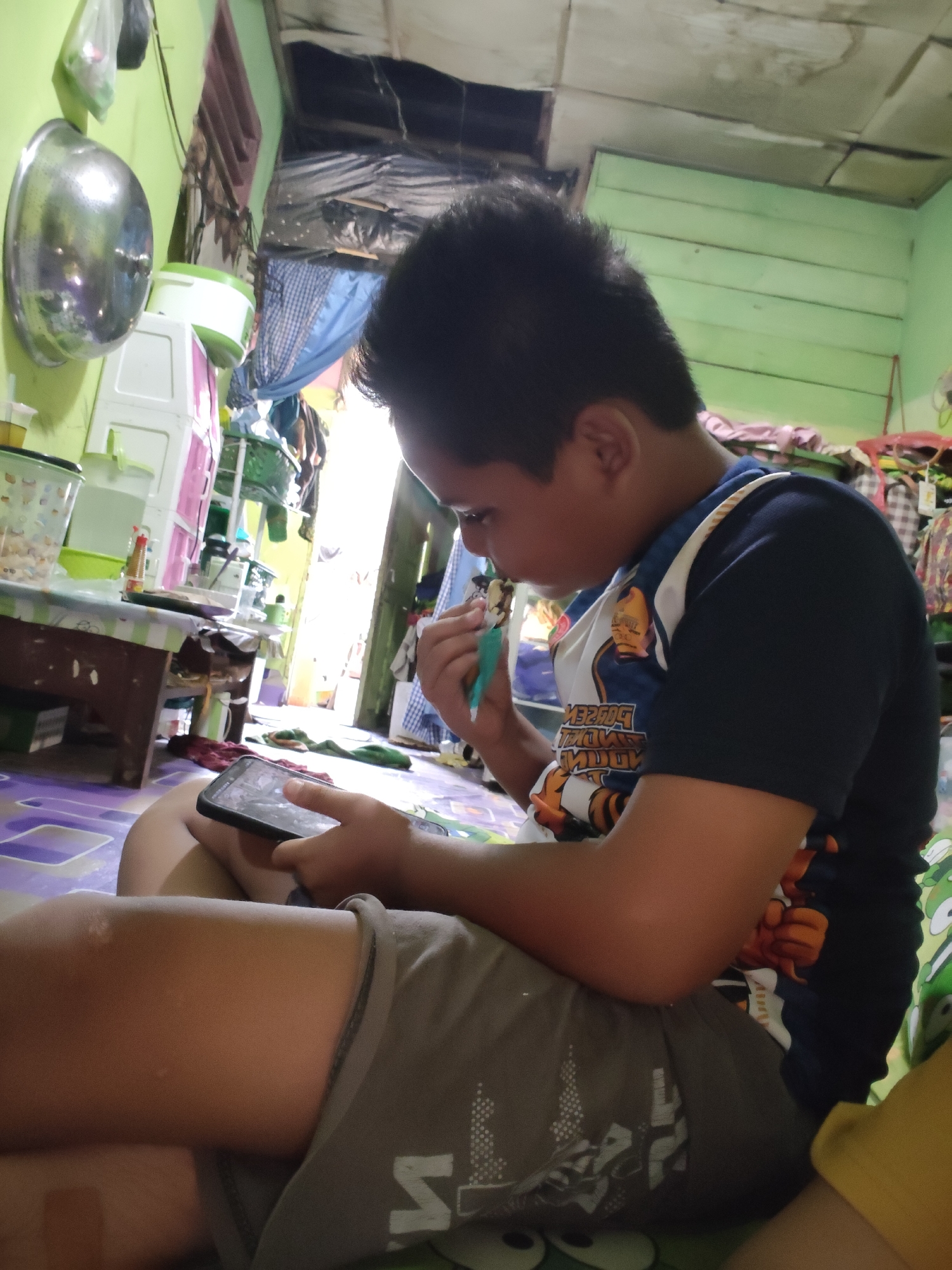 In a dimly lit room in Lori, Indonesia, a young boy sits on the floor, engrossed in his smartphone. He wears a dark blue t-shirt with a graphic print and grey shorts. His left hand holds the phone, its screen illuminated and reflecting in his eyes. In his right hand, he holds a small, light-colored object, possibly a toy or a tool, and appears to be interacting with it. The room is cluttered with various items, including plastic containers, what seems to be a white cabinet with pink drawers, and clothes hanging in the background. A metal bowl is mounted on the wall to the left, and a patterned mat covers the floor. The overall impression is of a humble, lived-in space.