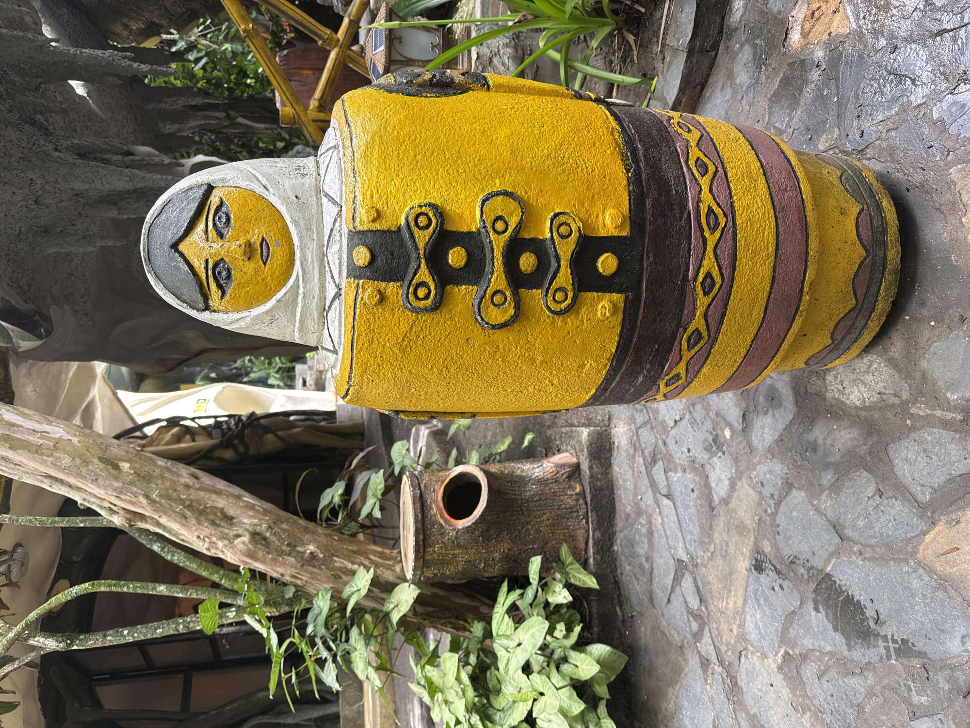 A brightly colored, large, cylindrical statue, reminiscent of a stylized doll or jar, dominates the outdoor scene. The statue is primarily painted in a vibrant yellow, with its upper portion featuring a face framed by a white hood or headscarf. The face itself is yellow with carved or painted black eyes, nose, and mouth, giving it a calm expression. Around the statue's mid-section, there's a broad black band adorned with three ornate, interconnected yellow decorative elements resembling buckles or clasps. Below this, the statue's body is decorated with horizontal bands of brown, yellow, and a patterned design featuring repeating diamond or zigzag motifs.
The statue rests on a ground covered with irregular grey flagstone paving. To its left, a gnarled, thick tree trunk extends horizontally, with smaller branches trailing down, covered in lush green foliage, possibly a type of ivy or philodendron with heart-shaped leaves. Behind the statue's base, a decorative log-shaped object with a circular opening, possibly a planter or birdhouse, is visible, surrounded by more greenery. In the background, hints of other structures, possibly made of wood or bamboo, along with more diverse plant life and sections of white fabric (perhaps a canopy or tent), suggest a landscaped environment.
The scene depicts a static, decorative display within what appears to be a garden or outdoor exhibition area. The lighting suggests it is daytime, possibly under an overcast sky or in a shaded spot, as there are no harsh shadows. There is no discernible activity or event taking place. The overall setting, characterized by the unique statue, natural stone path, and abundant greenery, aligns with the typical aesthetics of scenic or recreational areas found in places like Da Lat, Vietnam. No visible text is present in the image.