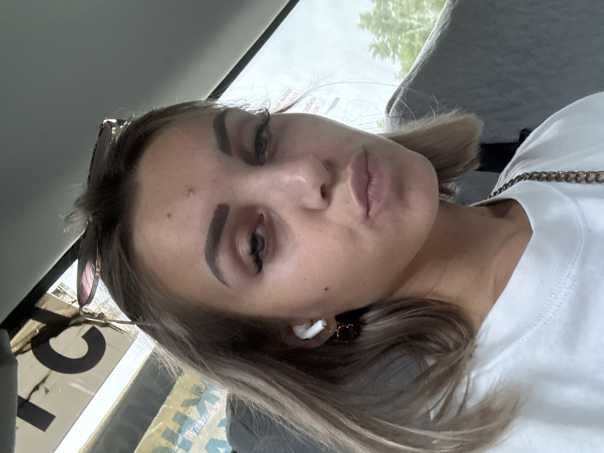 A young woman with blonde hair and makeup is captured in a close-up selfie, likely taken from a vehicle. She is wearing a white t-shirt and has an earbud in her left ear and a pair of sunglasses resting on her head. The background is blurred, but suggests an outdoor environment with trees visible through a window. Text is visible on the window, potentially indicating a location. The image appears to have been taken during the daytime.