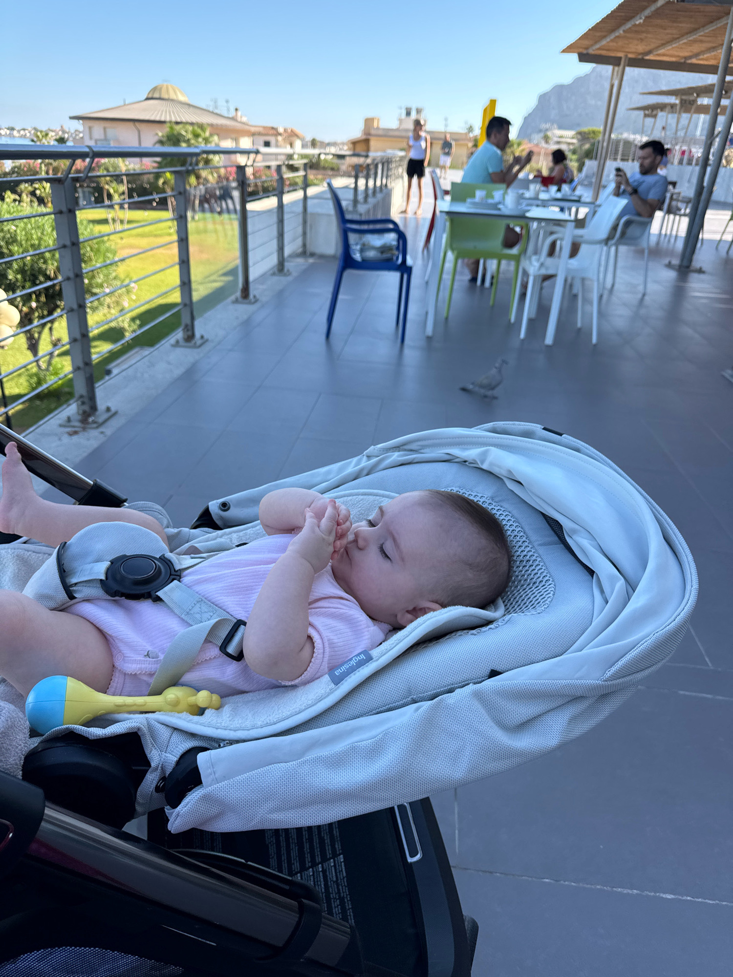 An outdoor dining area in Isola delle Femmine, Italy, is shown, featuring a baby asleep in a stroller in the foreground. The baby is dressed in a pink onesie and secured with a harness. A yellow and blue rattle lies next to the baby. In the background, people are seated at tables, some dining, others engaged with their phones. The setting appears to be a restaurant or cafe with a view of the sea and a building with a domed roof. A pigeon is visible on the pavement in the middle ground. The day is sunny with a clear blue sky.