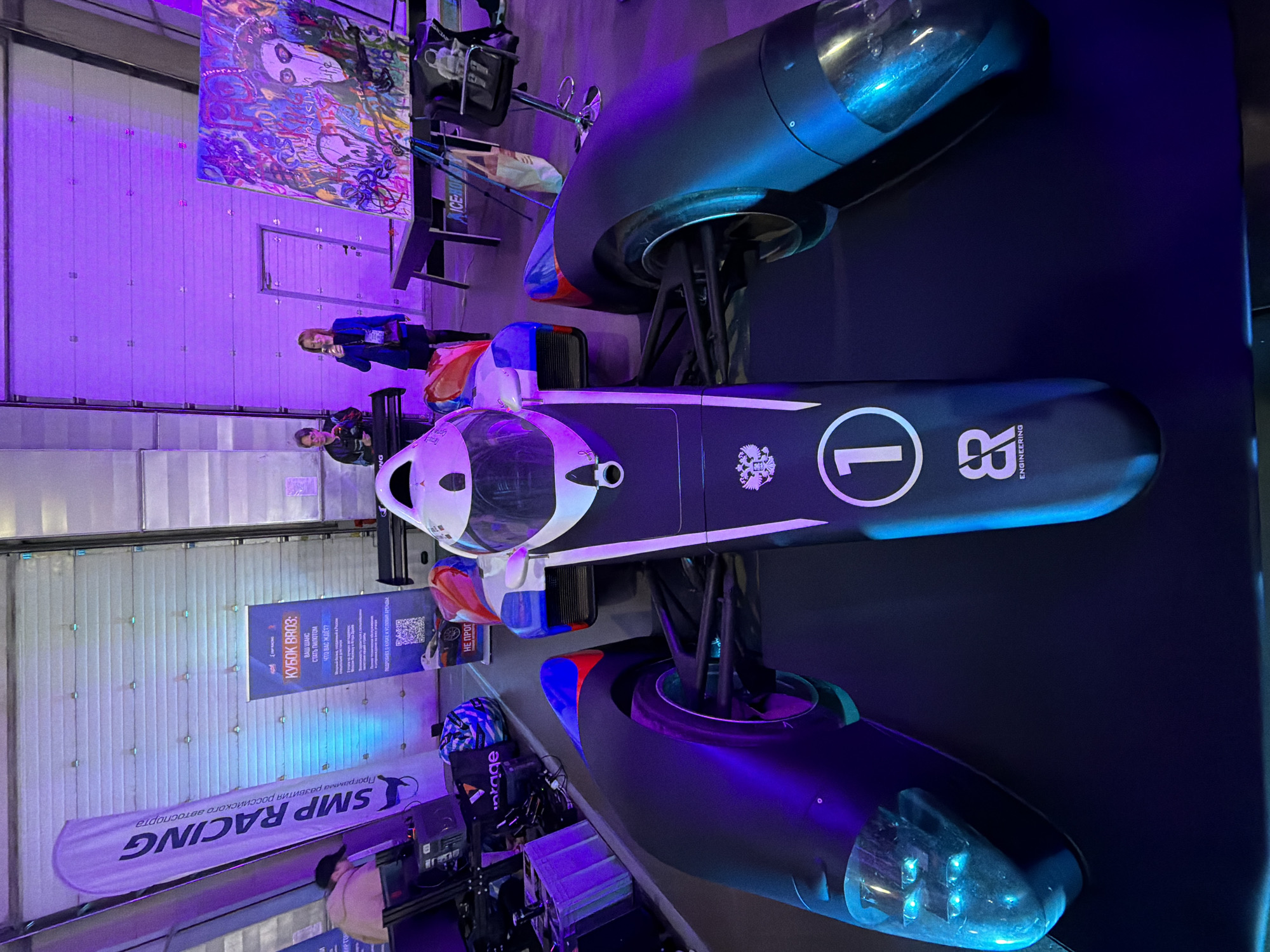 The image shows a tilted overhead view of a futuristic racing vehicle with prominent pontoons, displayed at what appears to be an indoor event. The vehicle is dark blue with white accents, and it features a large number "1" on the top surface, along with "18 Engineering" graphics. The pontoons are dark with clear lights at the front.
In the background, there is an assortment of items including banners, signs, and what may be racing-related equipment. A banner labeled "SMP RACING" and another banner with Russian text are visible. Two people are standing near a wall; one appears to be wearing a blue jacket. Behind them is an abstract painting and a display rack holding miscellaneous objects.
The scene is illuminated with purple and blue lighting, creating a colorful atmosphere. The overall setting suggests an automotive or racing exhibition, possibly located in Khimki, Russia, judging by the presence of the Russian text on the banners.