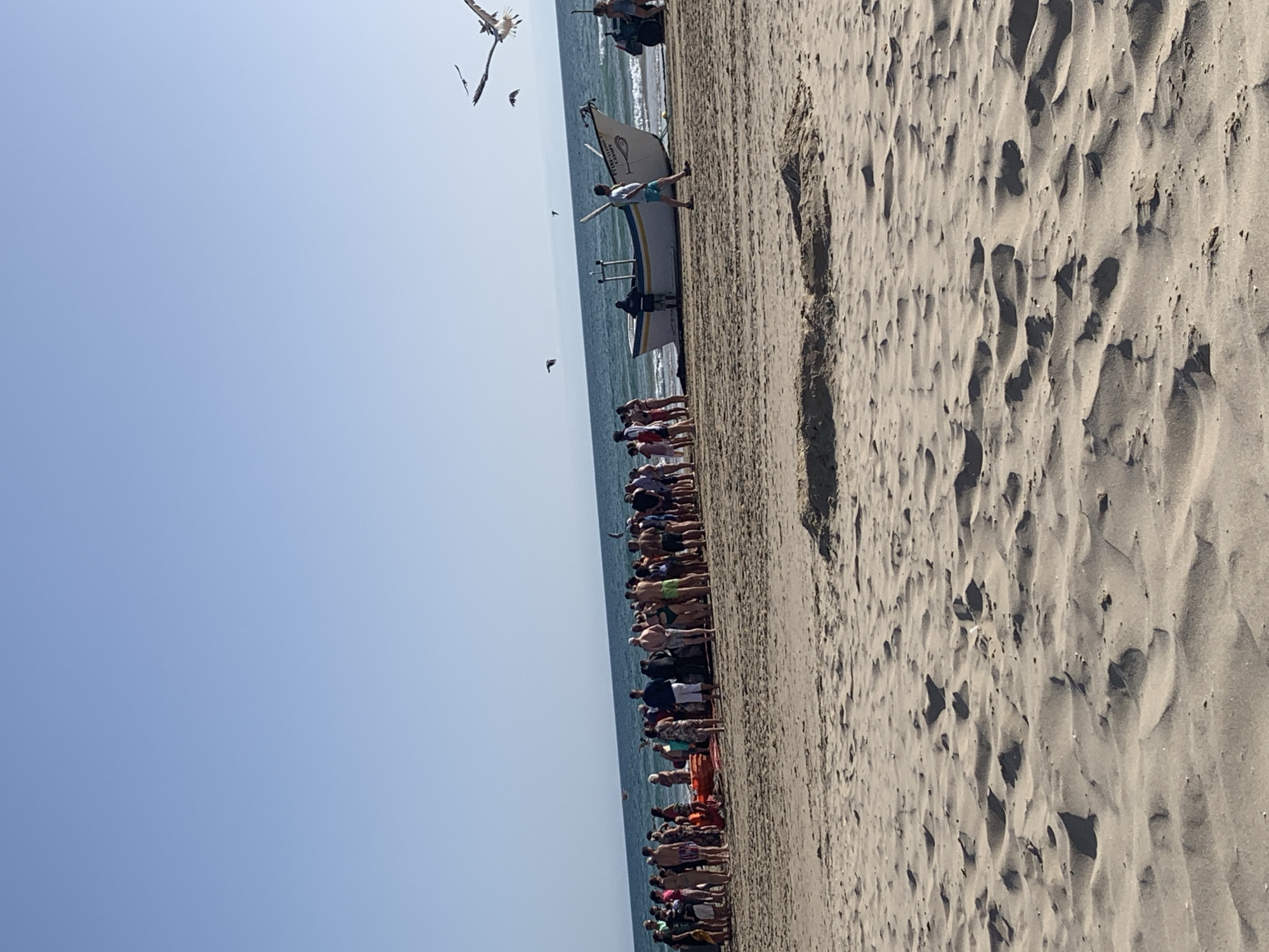 In Costa da Caparica, Portugal, a group of people stand on a sandy beach near the water's edge. Several individuals are gathered around a small, white fishing boat with blue trim, which is partially beached on the sand. Some people are looking at the boat, while others appear to be socializing. A few seagulls are in flight in the clear blue sky. The ocean is visible in the background, with gentle waves lapping the shore. The sand dunes and the open expanse of the beach suggest a typical coastal environment.