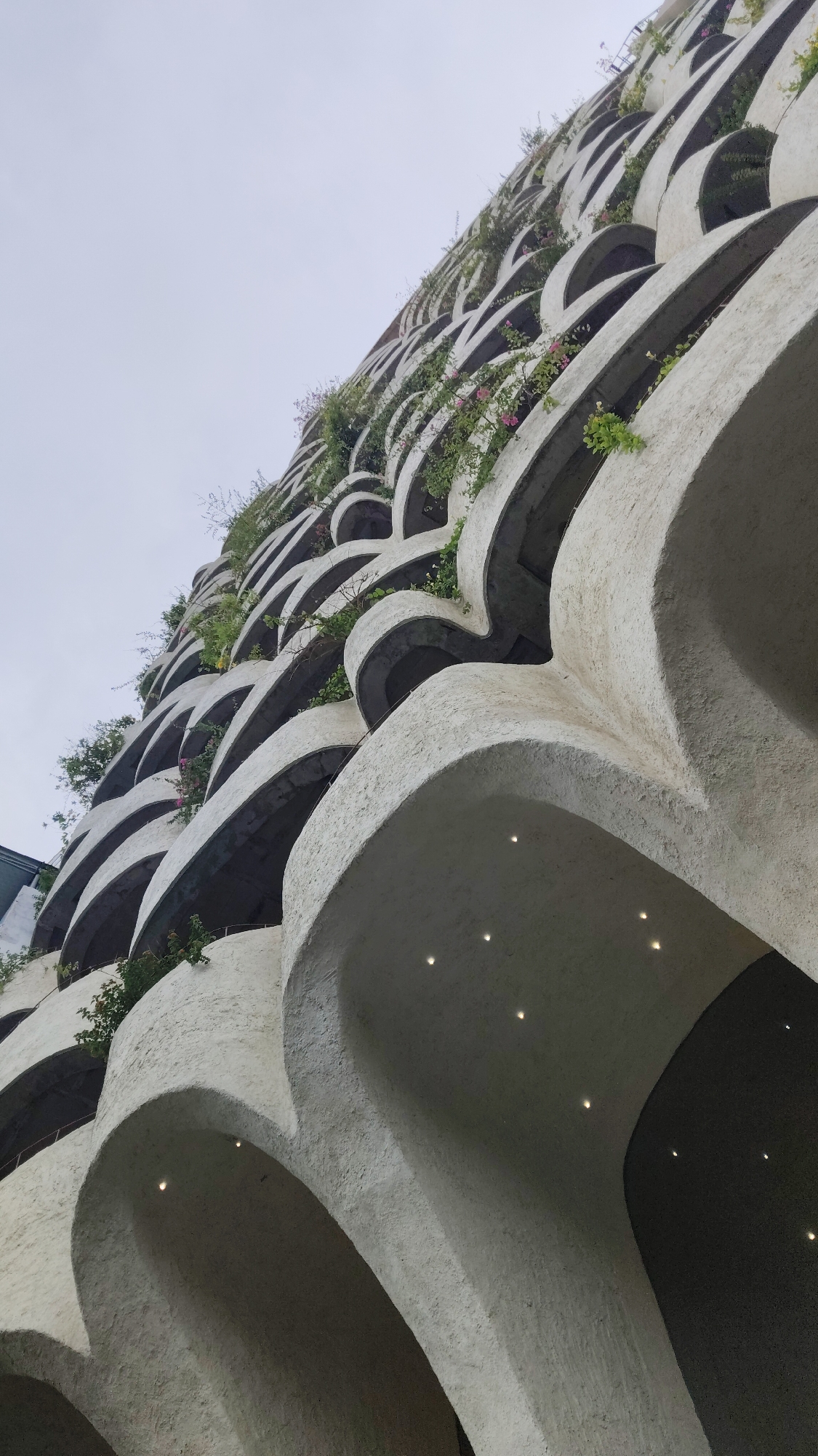 The image shows a close-up, low-angle view of a building's facade with distinctive architectural features. The building is characterized by a series of semi-circular, arched balconies stacked vertically, creating a textured, wave-like effect. The exterior of the building appears to be constructed from a light-colored, rough material, possibly concrete or stucco. The lighting underneath some of the balconies reveals small, recessed lights. Various plants and greenery are visible growing from the balconies. The image was taken in Bac Nha Trang Ward, Vietnam.
