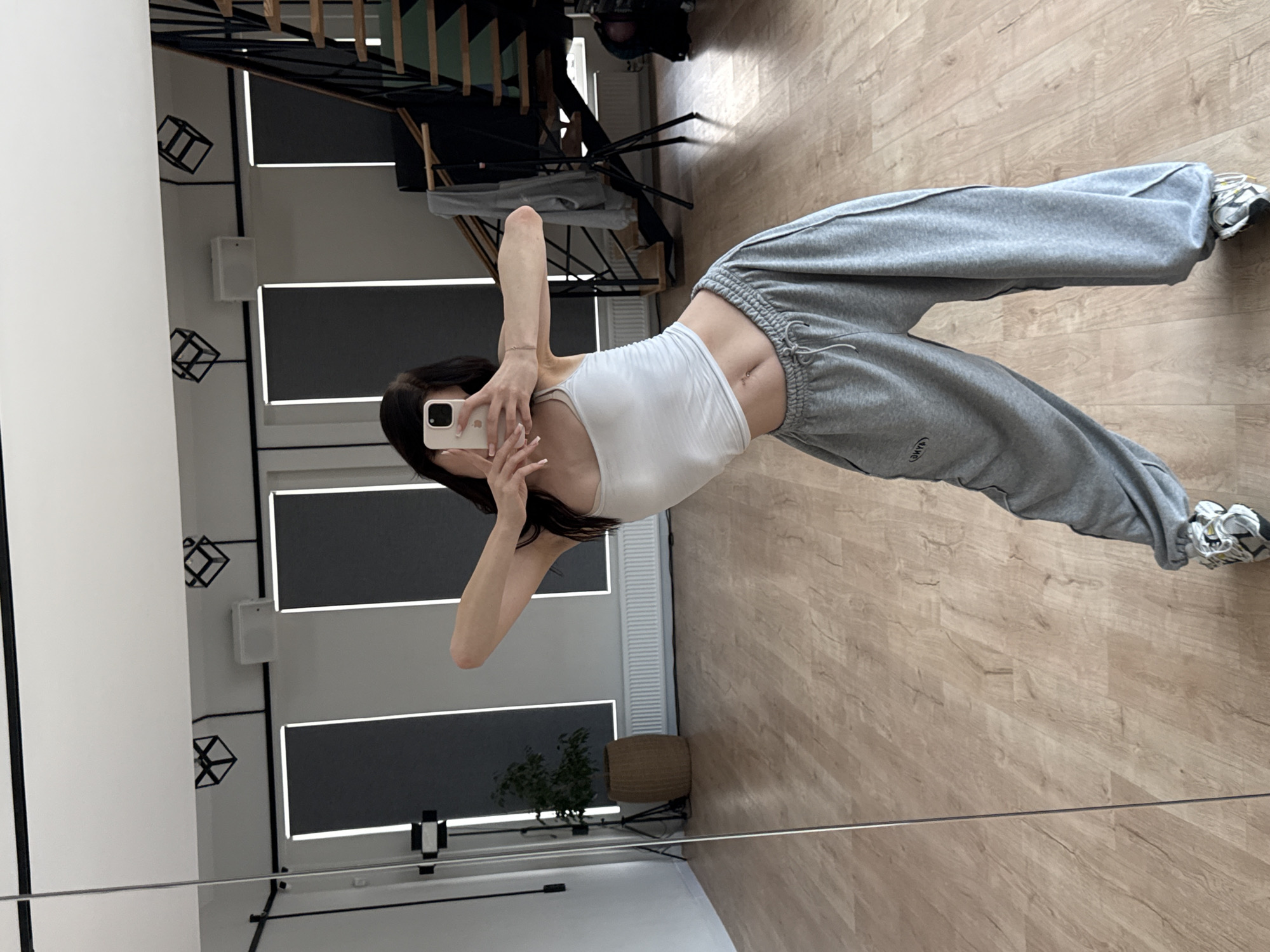 The image shows a young woman in a dance studio, possibly located in Agalarov Estate, Russia. She is wearing a white tank top, grey sweatpants, and sneakers, and is taking a mirror selfie. The studio has a wooden floor, large mirrors, and minimalist decor with artistic light fixtures on the ceiling. There is a plant in a woven pot and a staircase with a railing visible in the background. The woman is in a dynamic pose, with her body arched and arms extended. The overall atmosphere is casual and athletic.