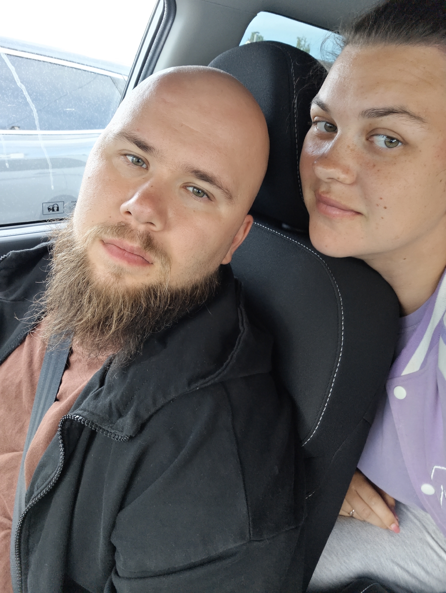 A selfie photograph depicts a man and a woman inside a vehicle. The man is bald with a full, light brown beard, wearing a black jacket and a brown shirt with a gray seatbelt crossing his chest. The woman, positioned to his right, has her hair pulled back and is wearing a lilac jacket with white accents. The car's headrest is black and white. Outside the car window is a gray car with a wet window. The location is Chaikovsky, Russia. No specific actions or interactions are taking place, other than posing for the photo.