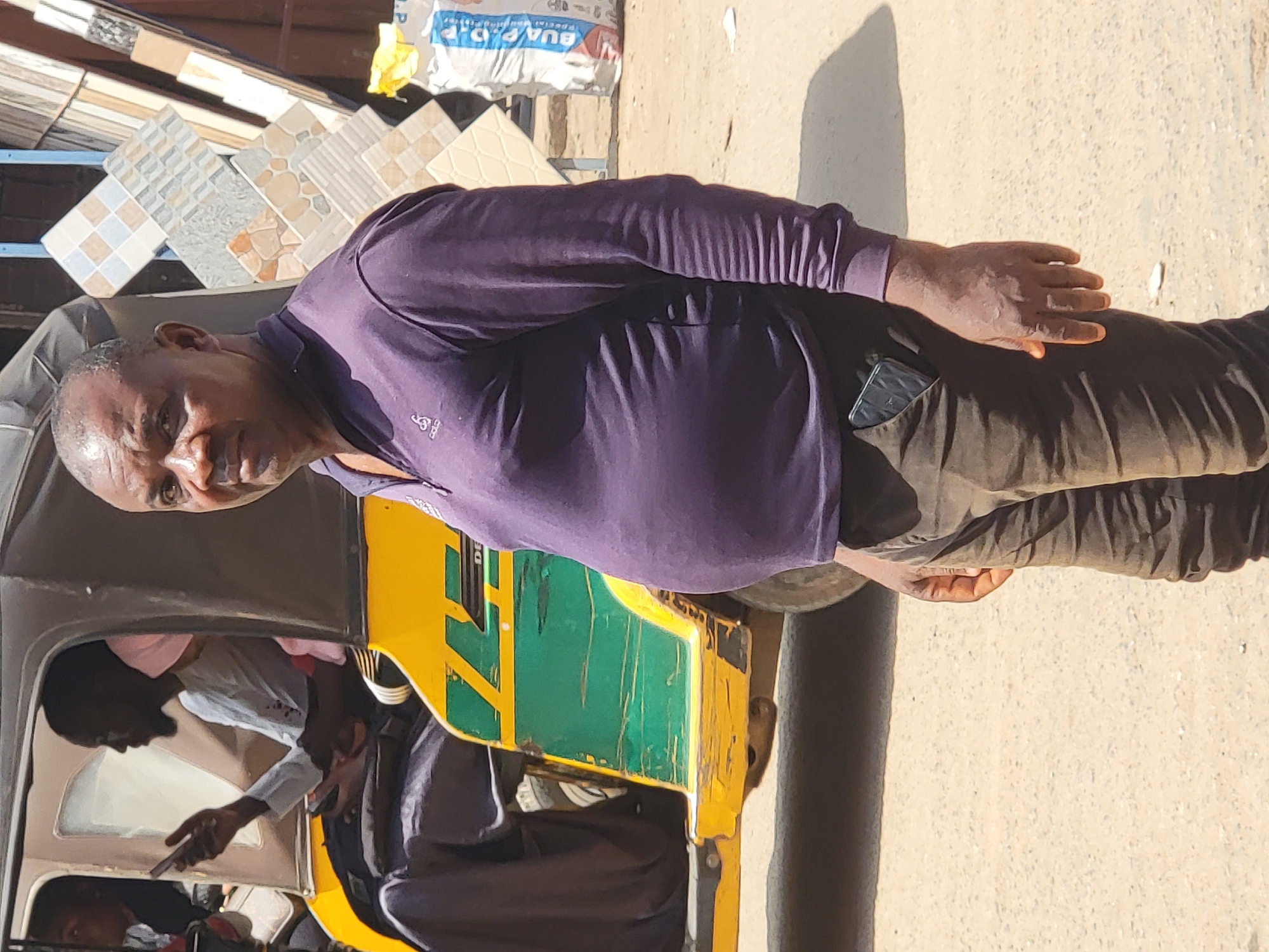 In Maiburiji, Nigeria, a man wearing a purple shirt and brown pants stands in front of a yellow and green auto rickshaw. There are passengers inside the auto rickshaw, and tiles stacked nearby on display. The ground is sandy and the sun is shining, casting shadows. The man has a phone in his pocket.